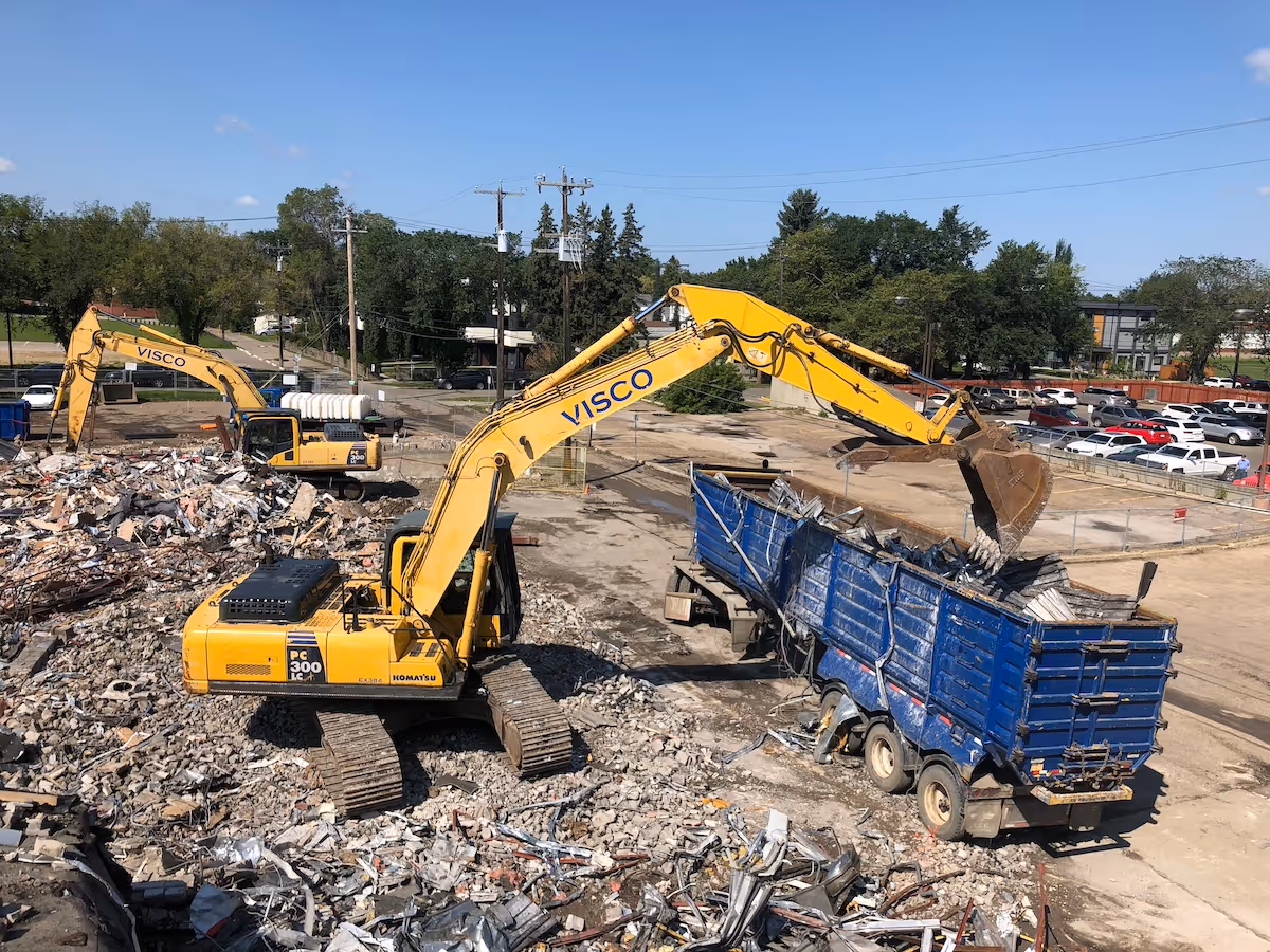 Yellow excavators loading debris into a blue dump truck at a construction demolition site under clear blue sky.