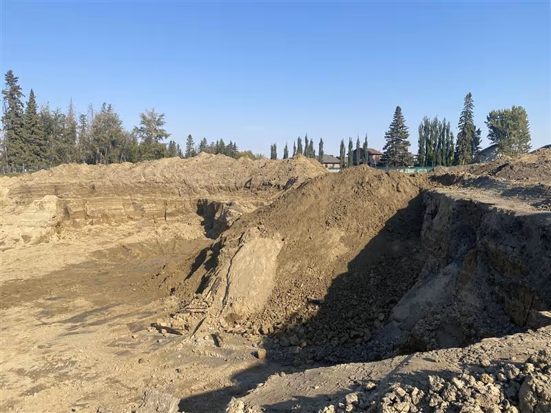Large excavation site with piles of dirt and soil against a background of trees and clear blue sky.