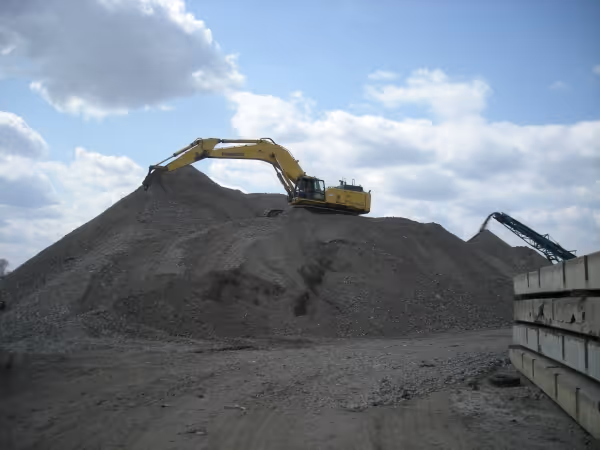 Yellow excavator on top of a large gravel pile at a construction or mining site under a cloudy blue sky.