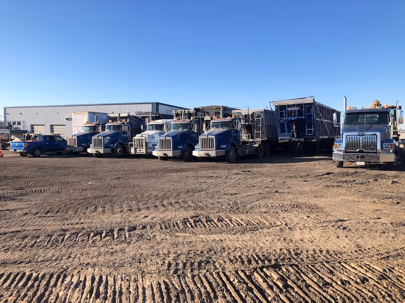 Row of blue heavy-duty trucks and trailers parked on dirt ground in front of industrial building under clear blue sky.