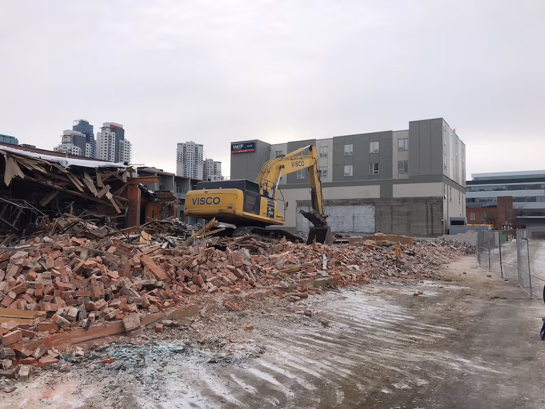 Yellow excavator labeled VISCO on a large pile of bricks and rubble at a demolition site with modern buildings in the background under a cloudy sky.