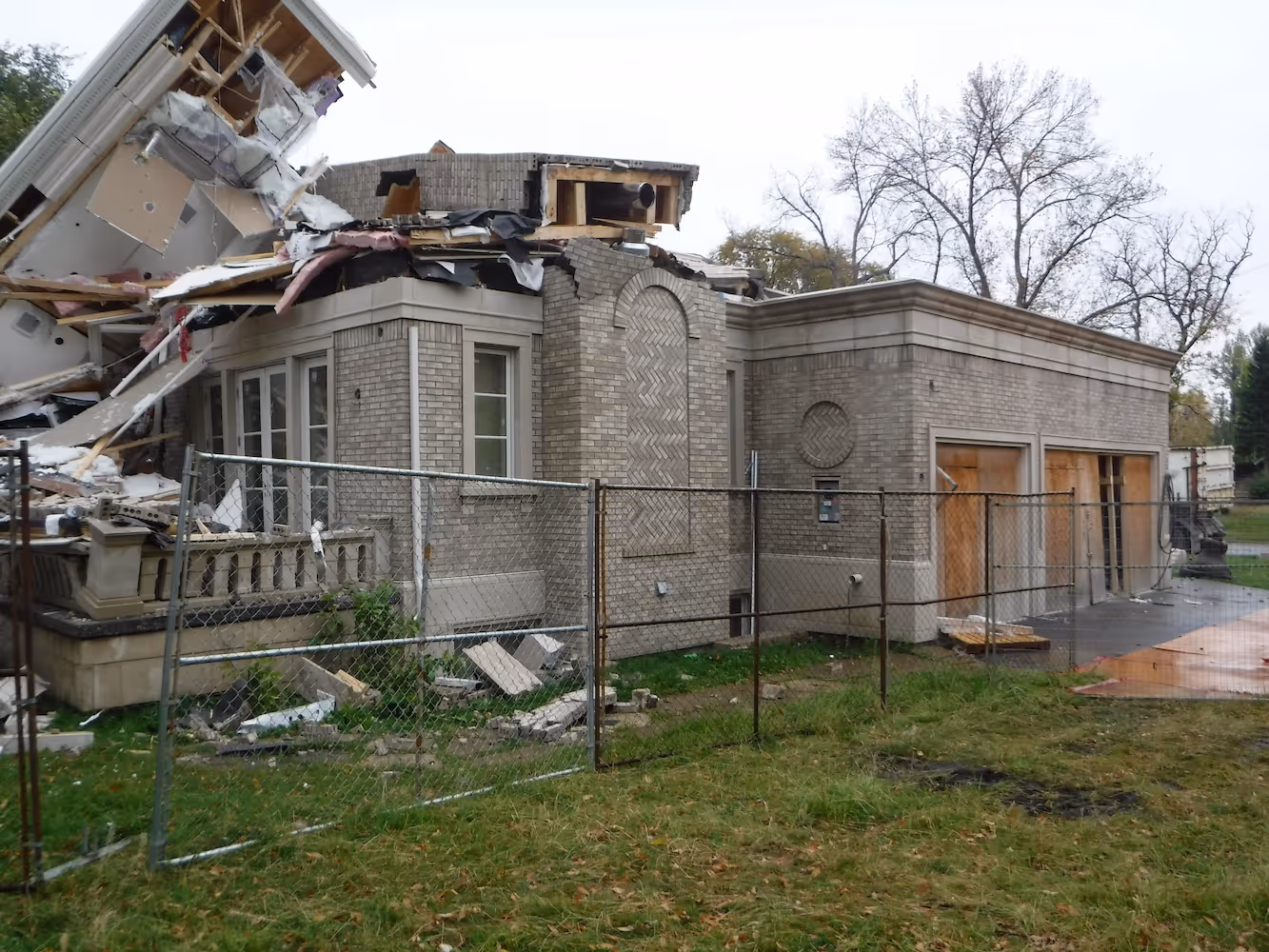 Partially collapsed brick house with extensive roof damage behind a chain-link fence on a grassy yard.