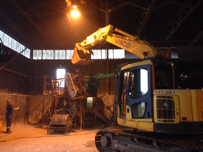 Yellow excavator with label VISCO operating inside a dimly lit industrial warehouse with a worker standing nearby.