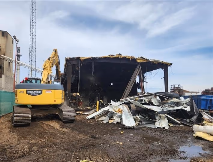 Yellow Komatsu excavator next to a partially demolished industrial building with debris pile on muddy ground.