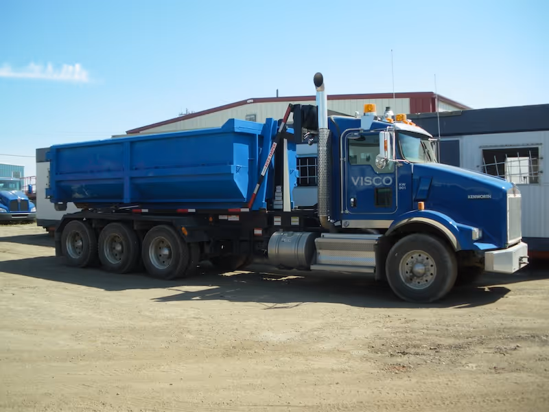 Blue Kenworth roll-off truck with a large blue container on a gravel surface near buildings.