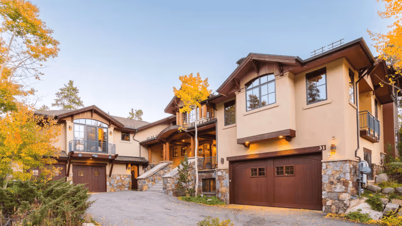 Large modern mountain home with beige stucco walls, stone accents, dark wood garage doors, balconies, and autumn trees around.