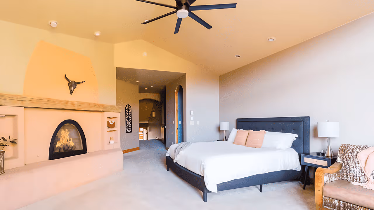 Spacious bedroom with light beige walls, a modern gas fireplace with a decorative bull skull above, a king-sized bed with pink pillows, nightstands with lamps, and an armchair with a blanket.