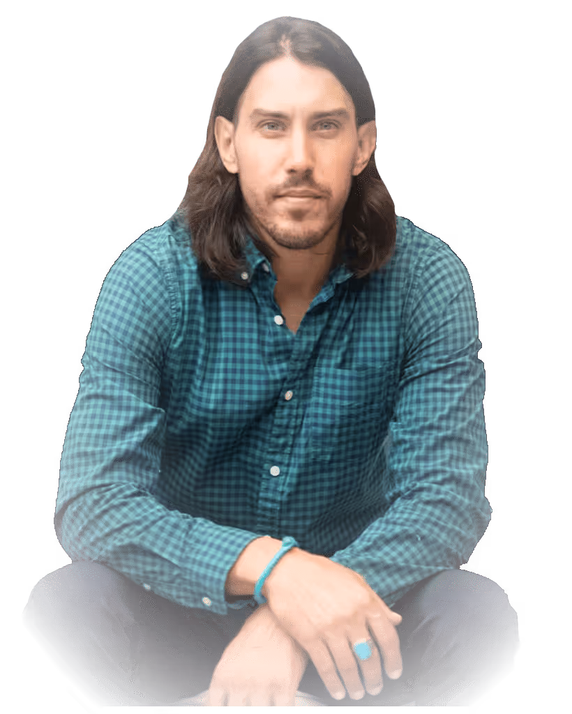 Man with long dark hair wearing a teal checkered shirt, sitting cross-legged and looking forward.