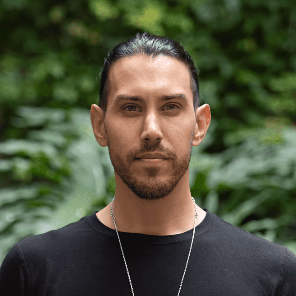 Portrait of a man with dark hair tied back, wearing a black shirt and silver necklace, with a leafy green background.