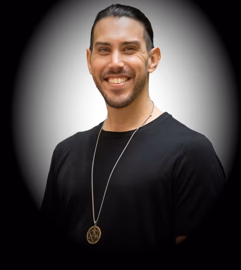 Smiling man with dark hair wearing a black shirt and a necklace with a round pendant.