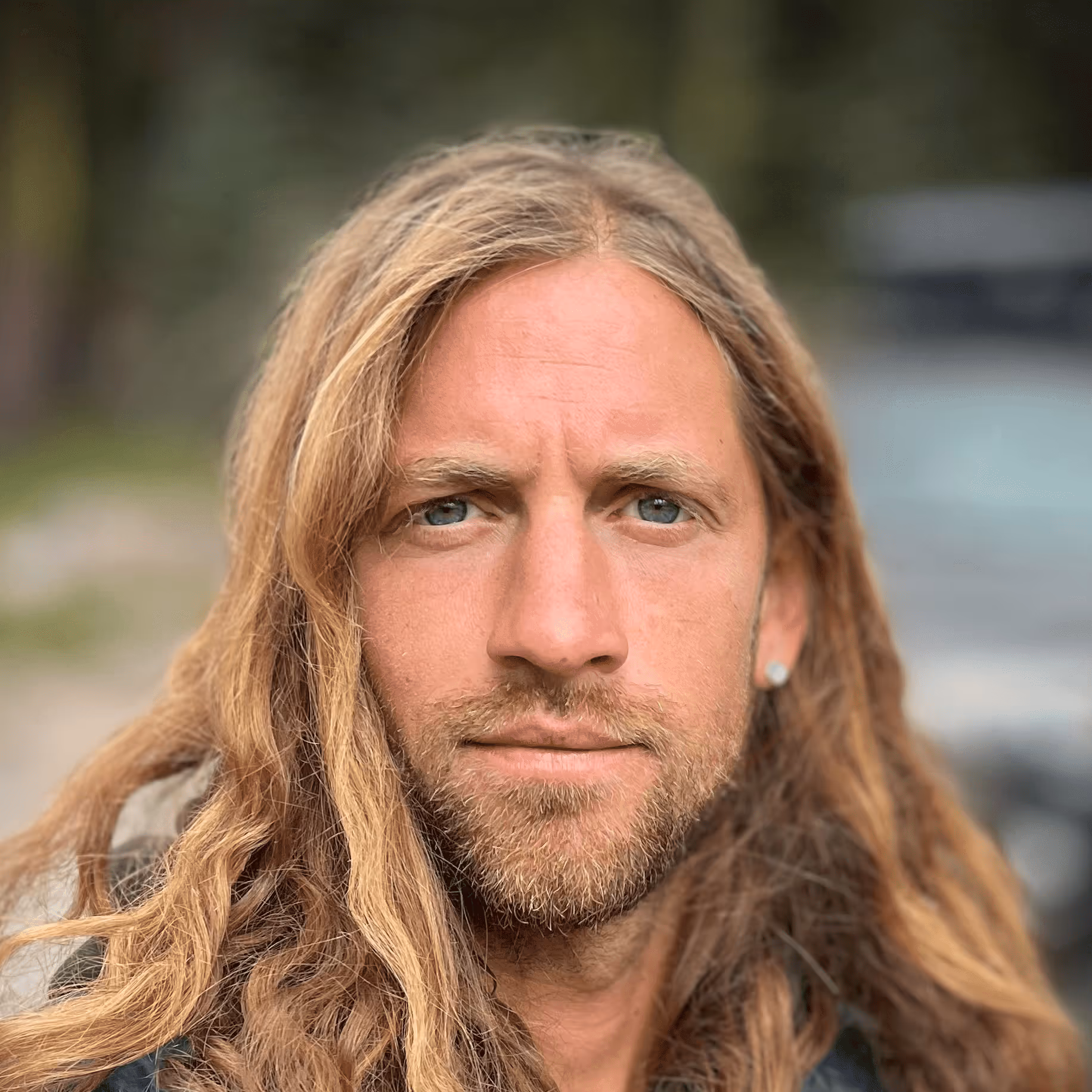 Close-up of a man with long blonde hair, blue eyes, and a short beard outdoors with a blurred background.