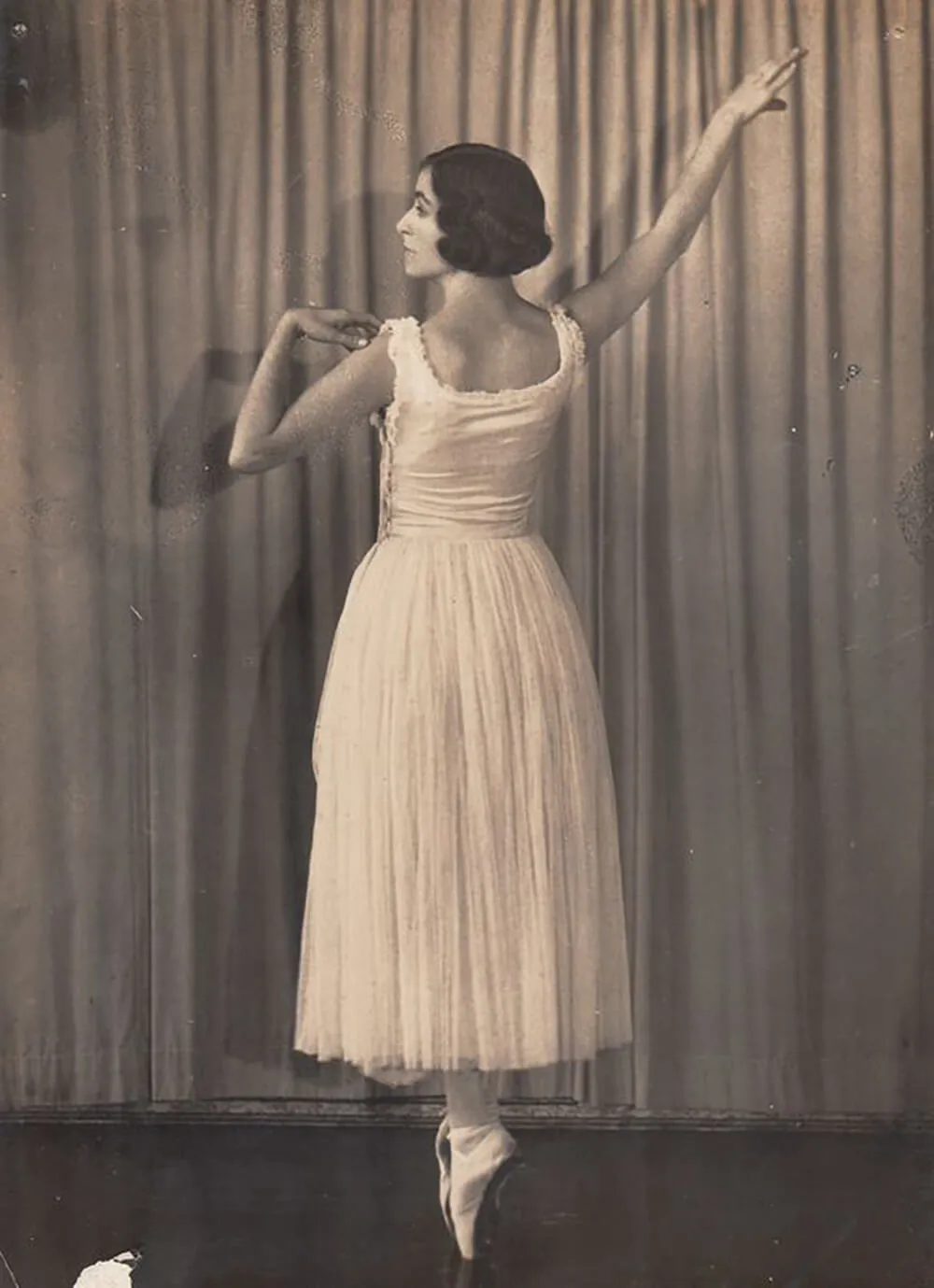 Vintage black-and-white photo of a female ballet dancer standing en pointe with arms gracefully raised, facing a curtain backdrop.