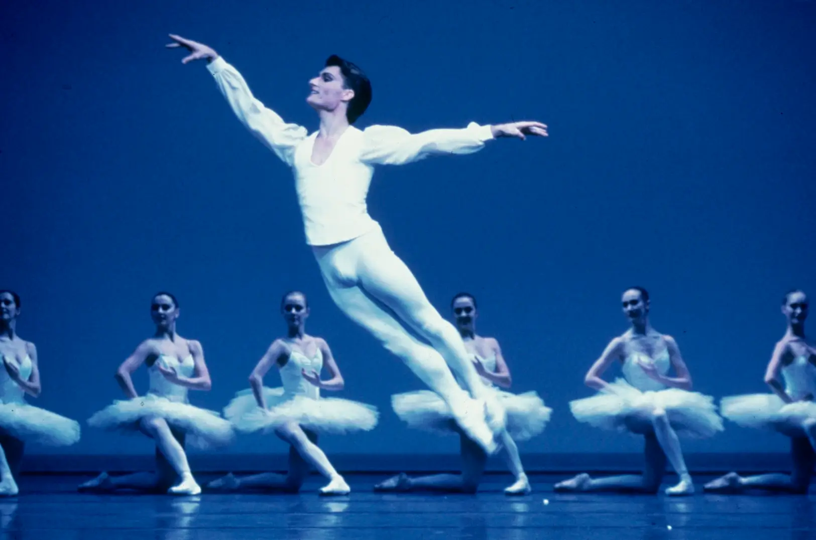 Male ballet dancer in white costume leaping in front of six female ballet dancers kneeling on stage in white tutus.