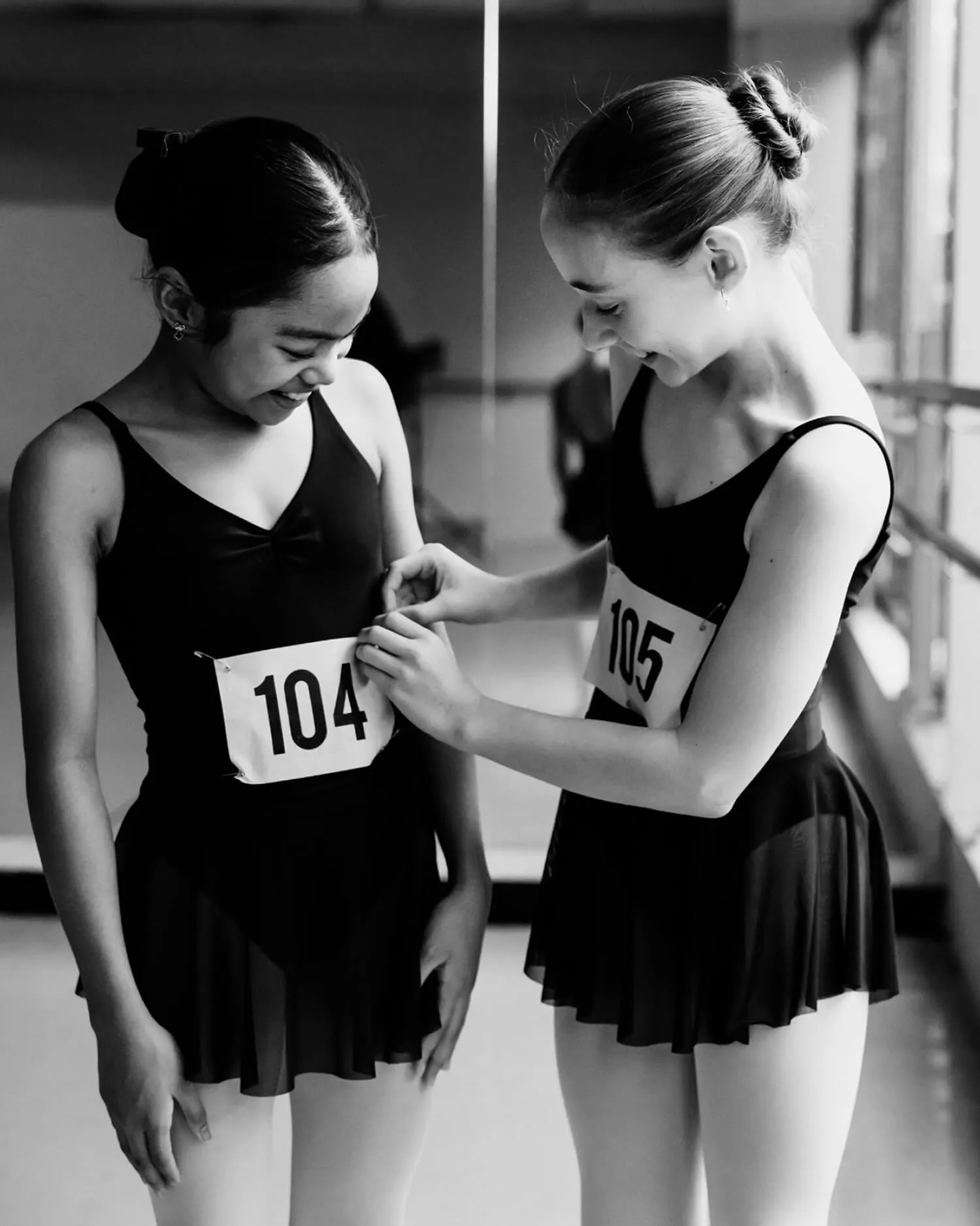 Two young ballerinas with numbered bibs smiling as one pins the other's bib in a dance studio.