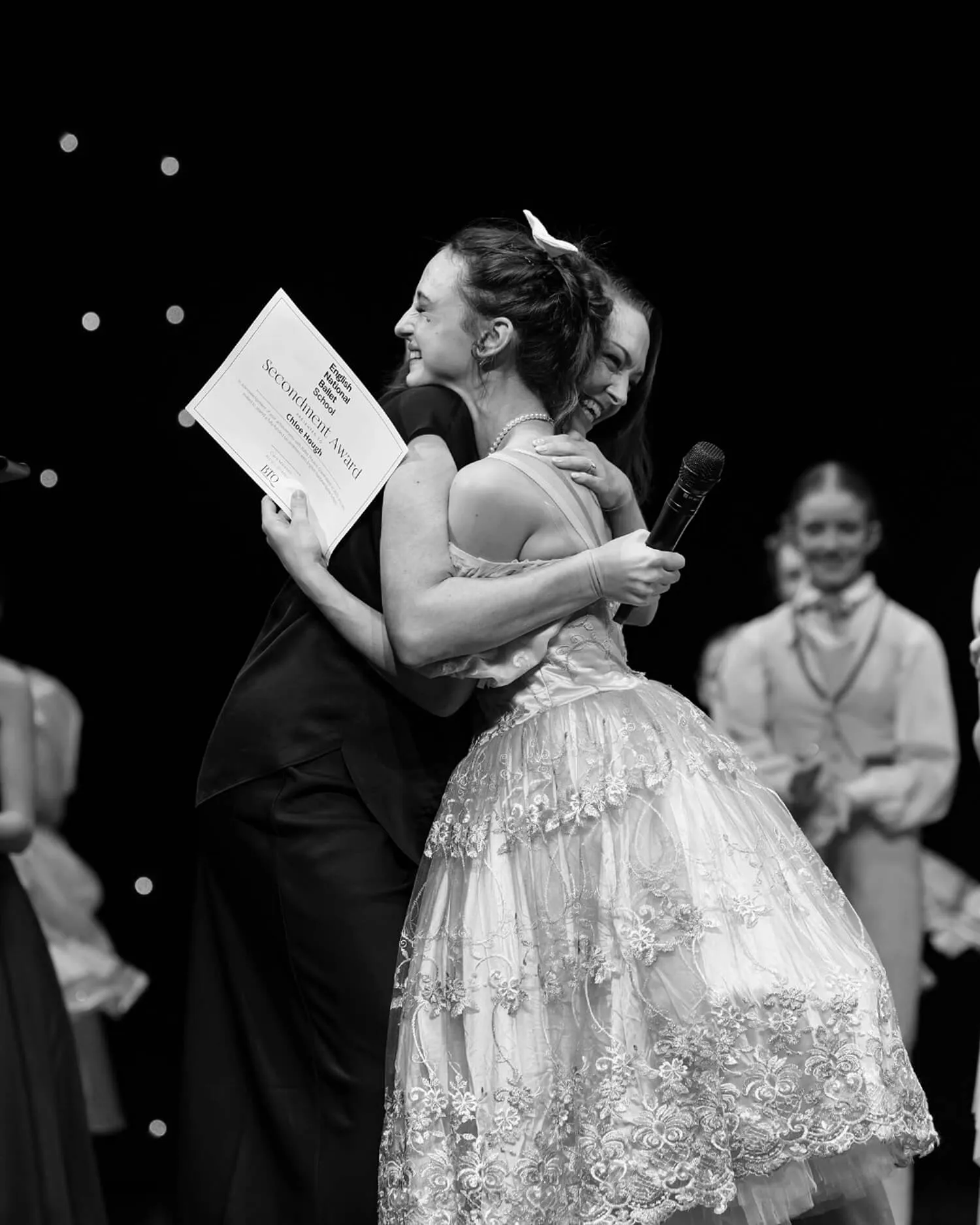 Two young women embrace happily on stage during an award ceremony, one holding a secondment award certificate and a microphone.
