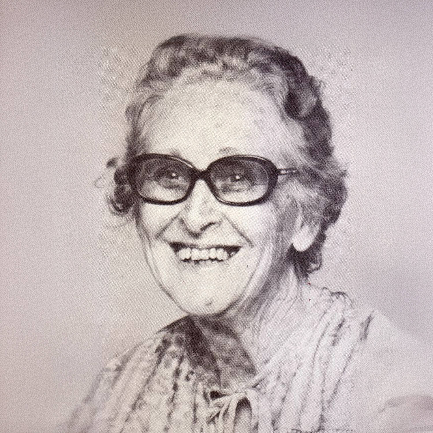 Smiling elderly woman with short curly hair wearing large dark glasses and a patterned blouse.