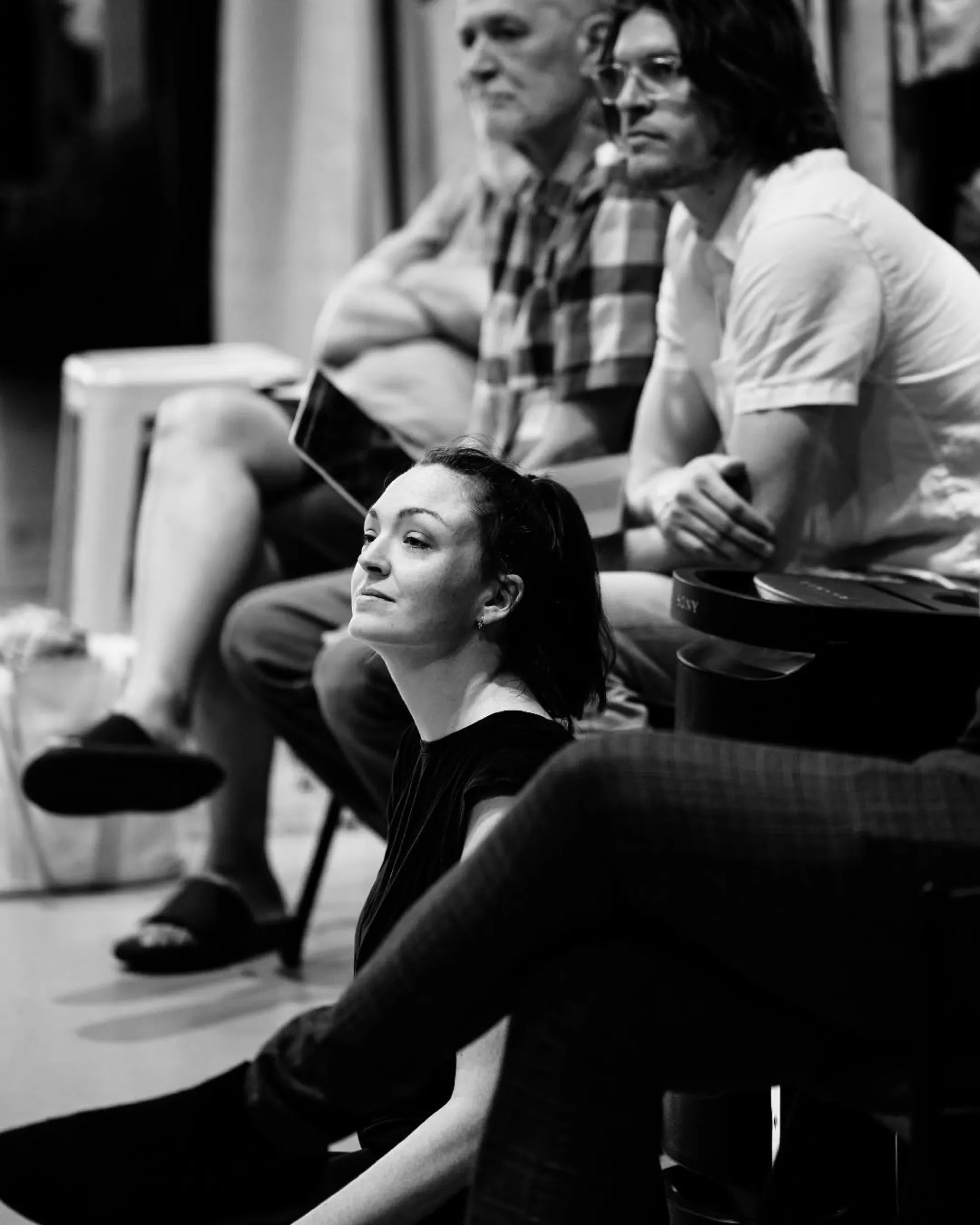 Black and white photo of a woman sitting on the floor looking thoughtfully ahead, with several people seated behind her.