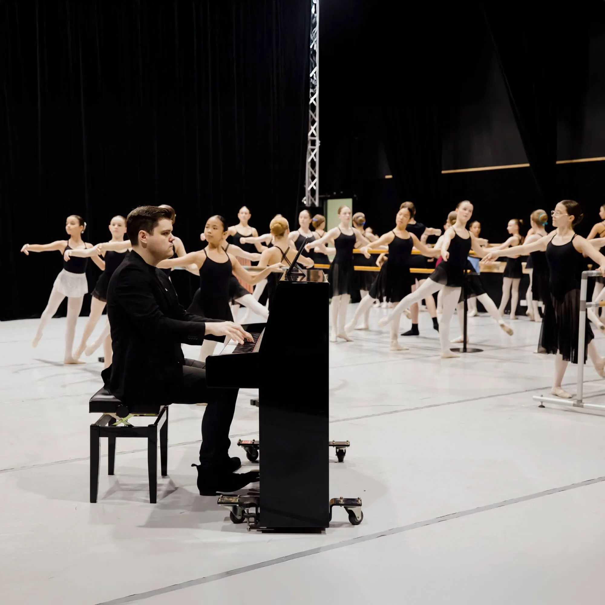 Man playing piano while ballet dancers practice at a barre in a dance studio.