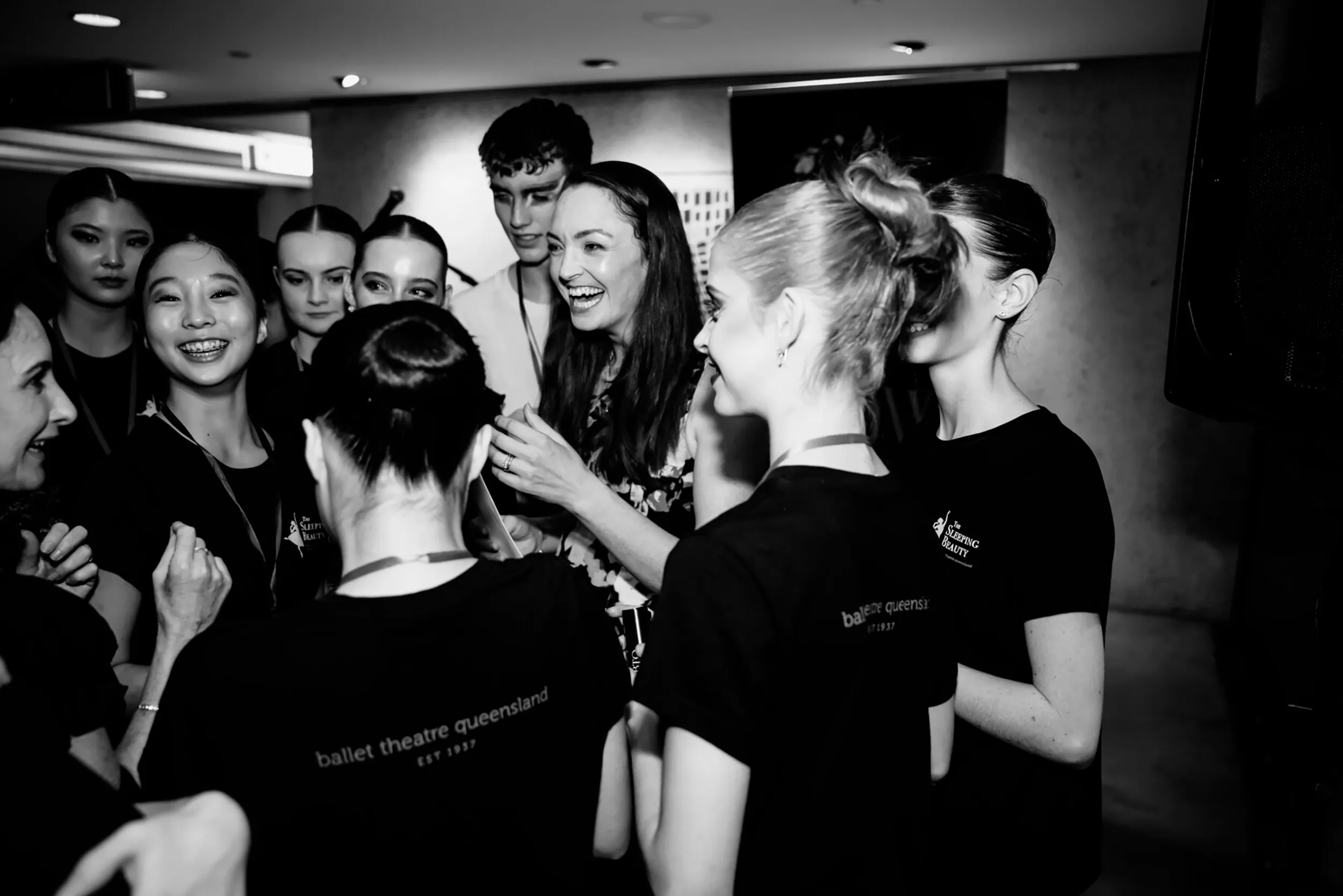 Group of ballet dancers and a smiling woman engaged in lively conversation backstage.