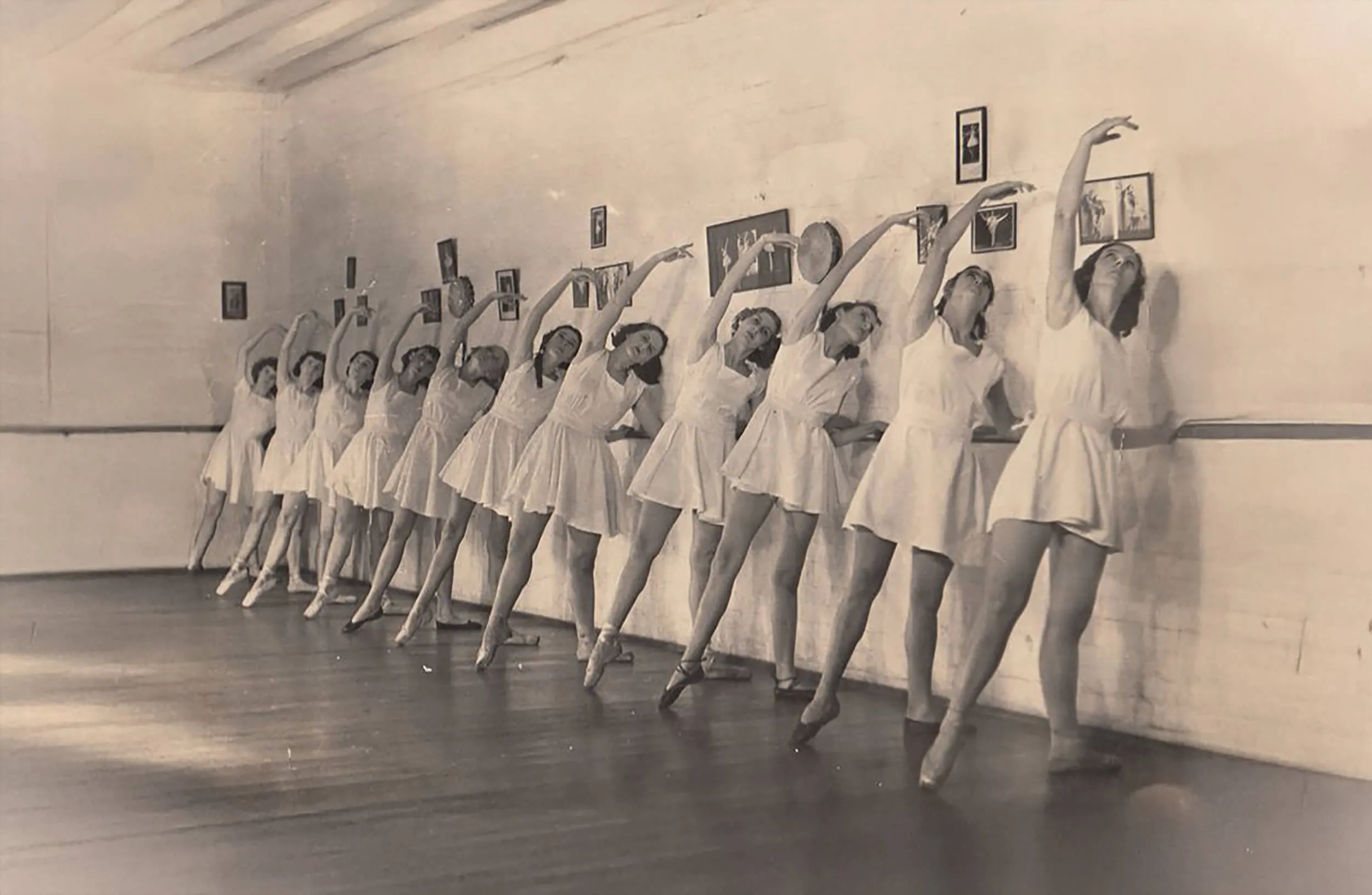 Black and white photo of a line of female ballet dancers in tutus practicing at a barre, all raising one arm gracefully.