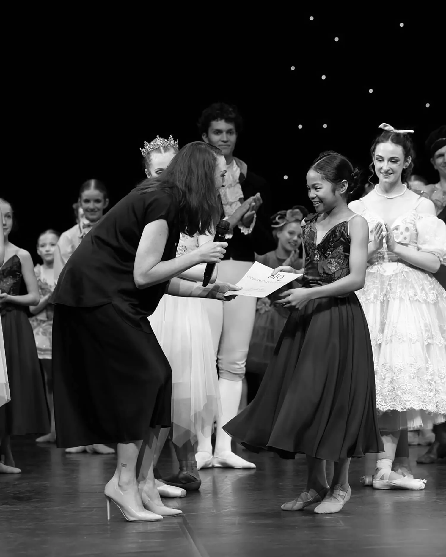Young ballerina receiving a certificate from a woman holding a microphone, with other costumed dancers clapping in the background.