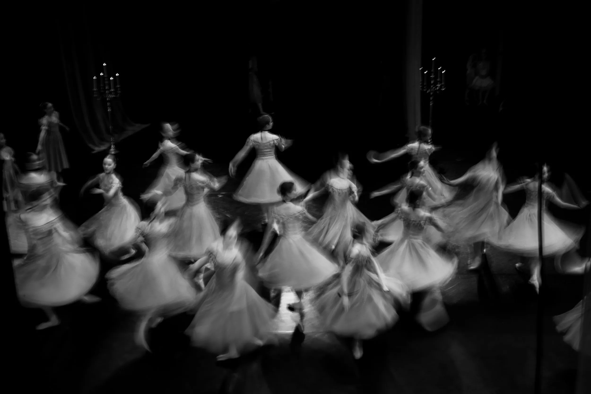 Black and white photo of ballerinas in flowing dresses dancing in a circle on stage.