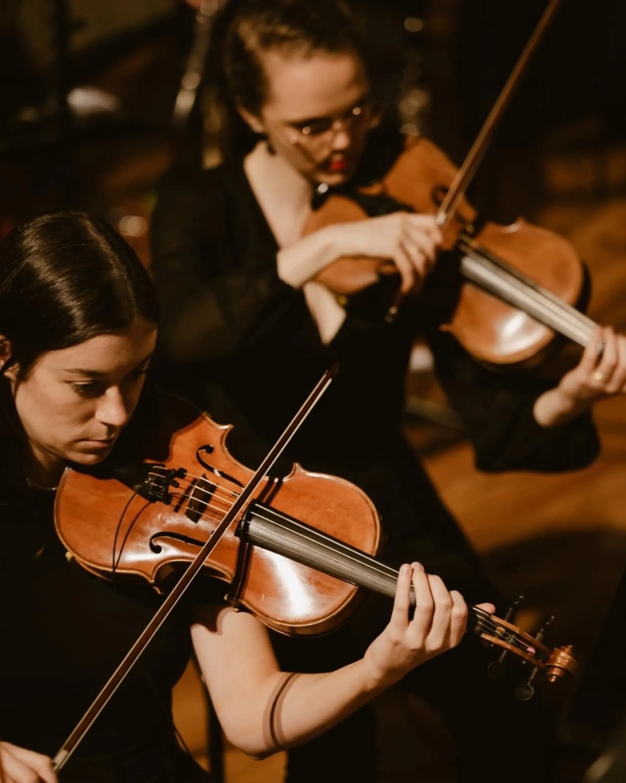 Two women playing violins in a dimly lit setting, focusing intently on their performance.