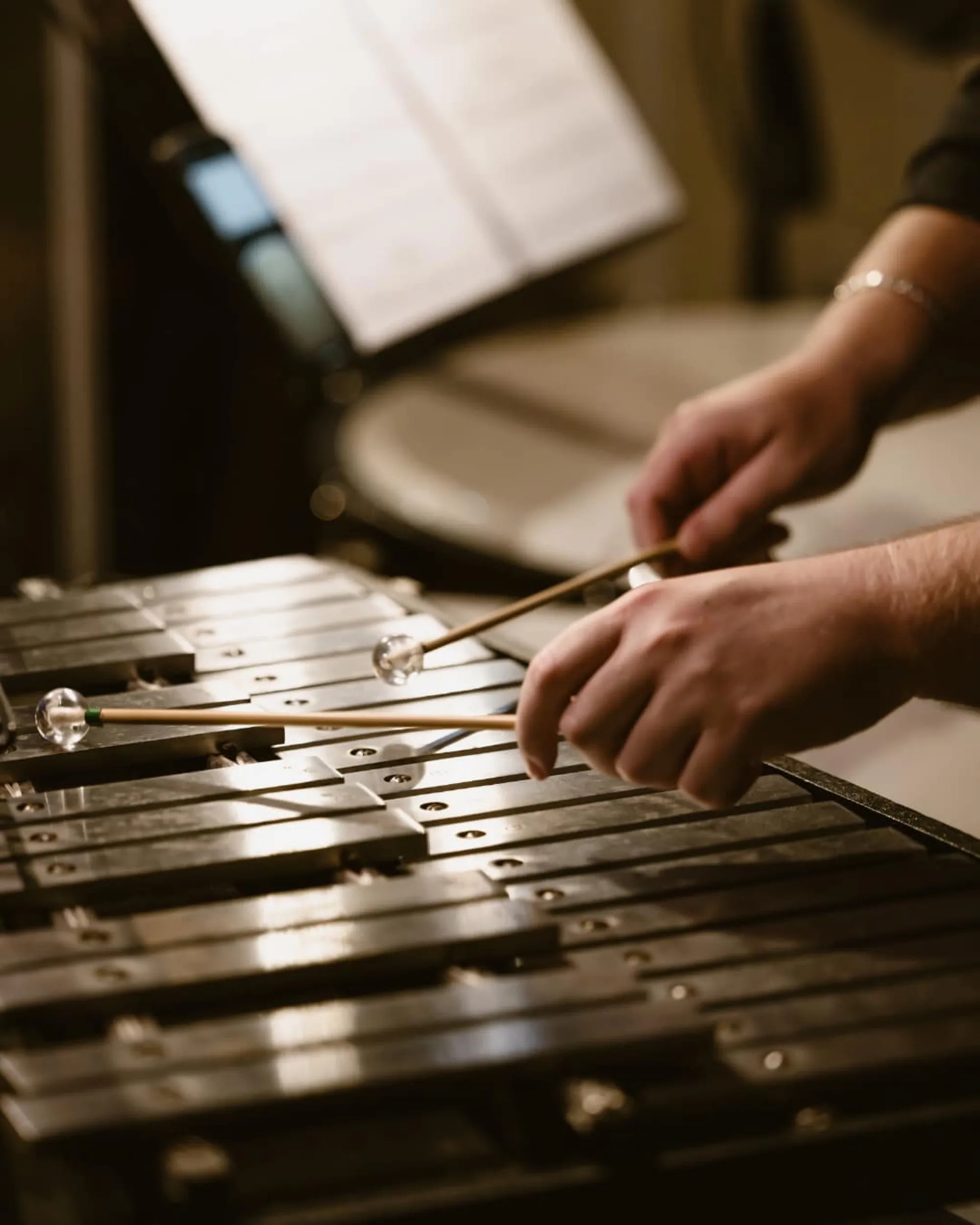 Hands playing a vibraphone with mallets, sheet music and a drum visible in the background.
