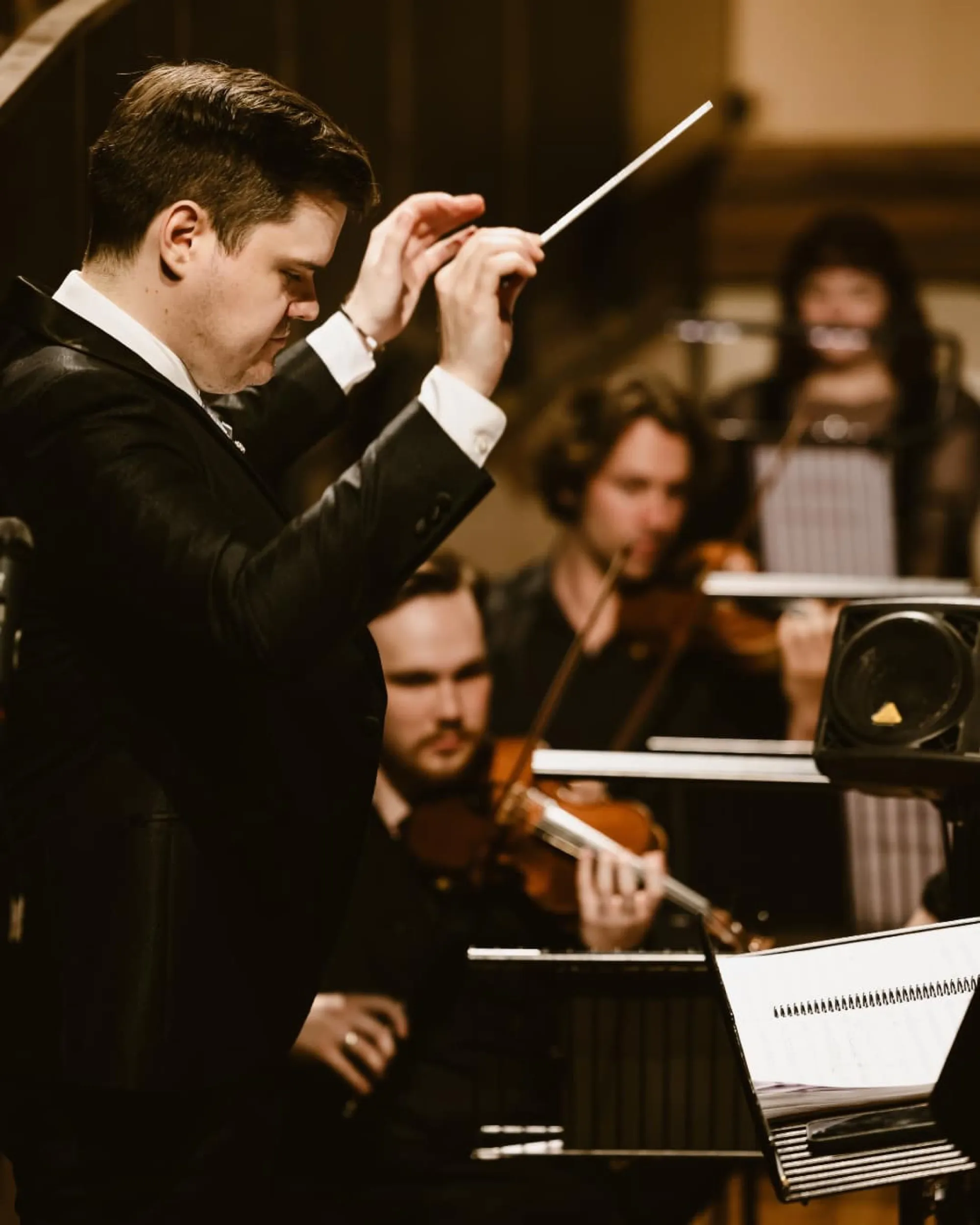 Orchestra conductor in black suit leading musicians playing violin and flute.