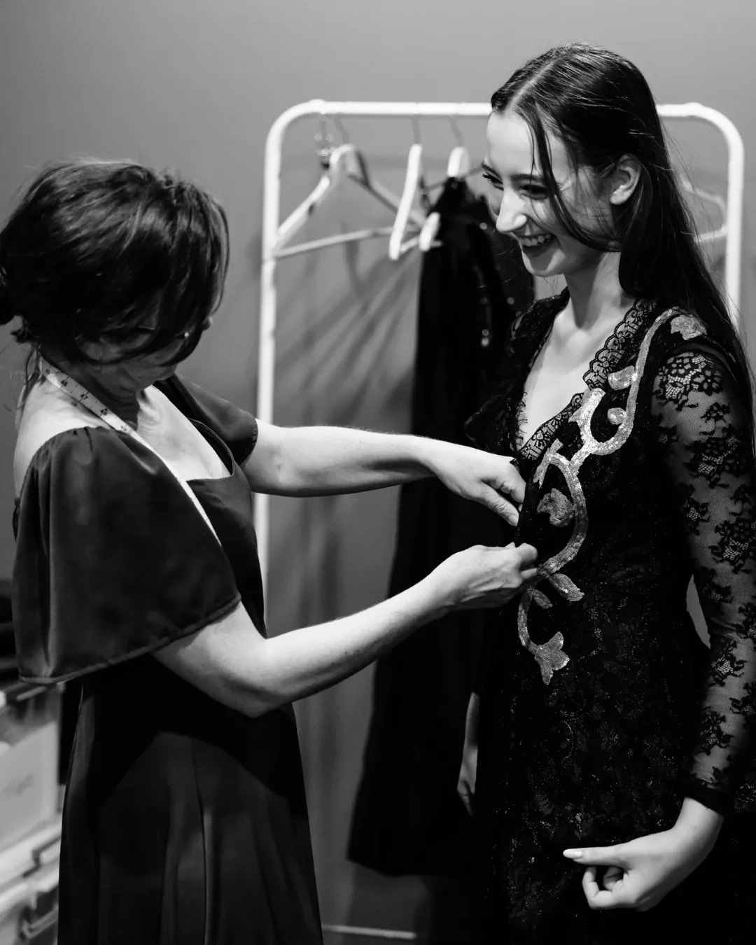 Seamstress adjusting a detailed lace dress on a smiling woman in a fitting room.