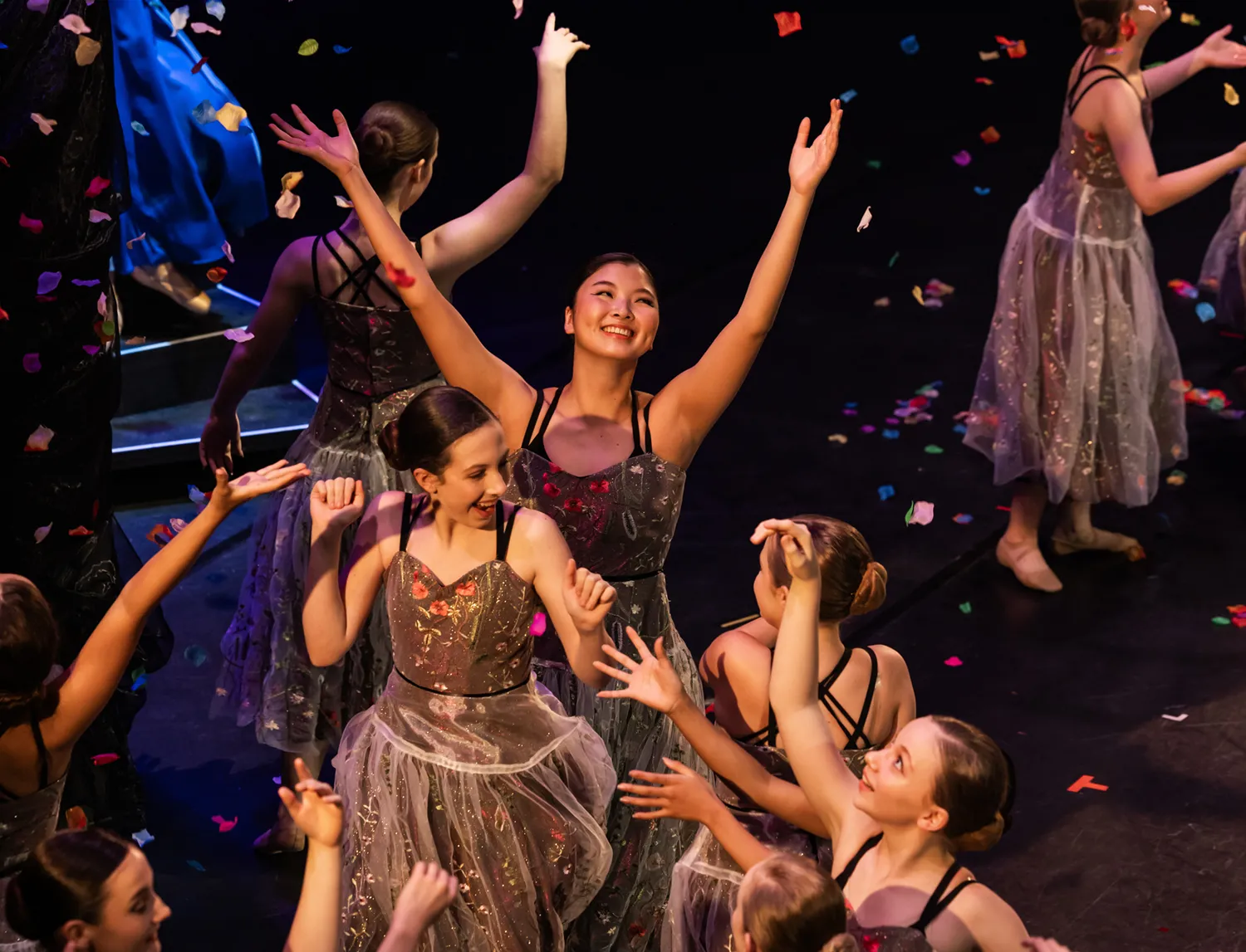 Group of joyful ballet dancers in sparkly dresses celebrating on stage with colorful confetti falling.