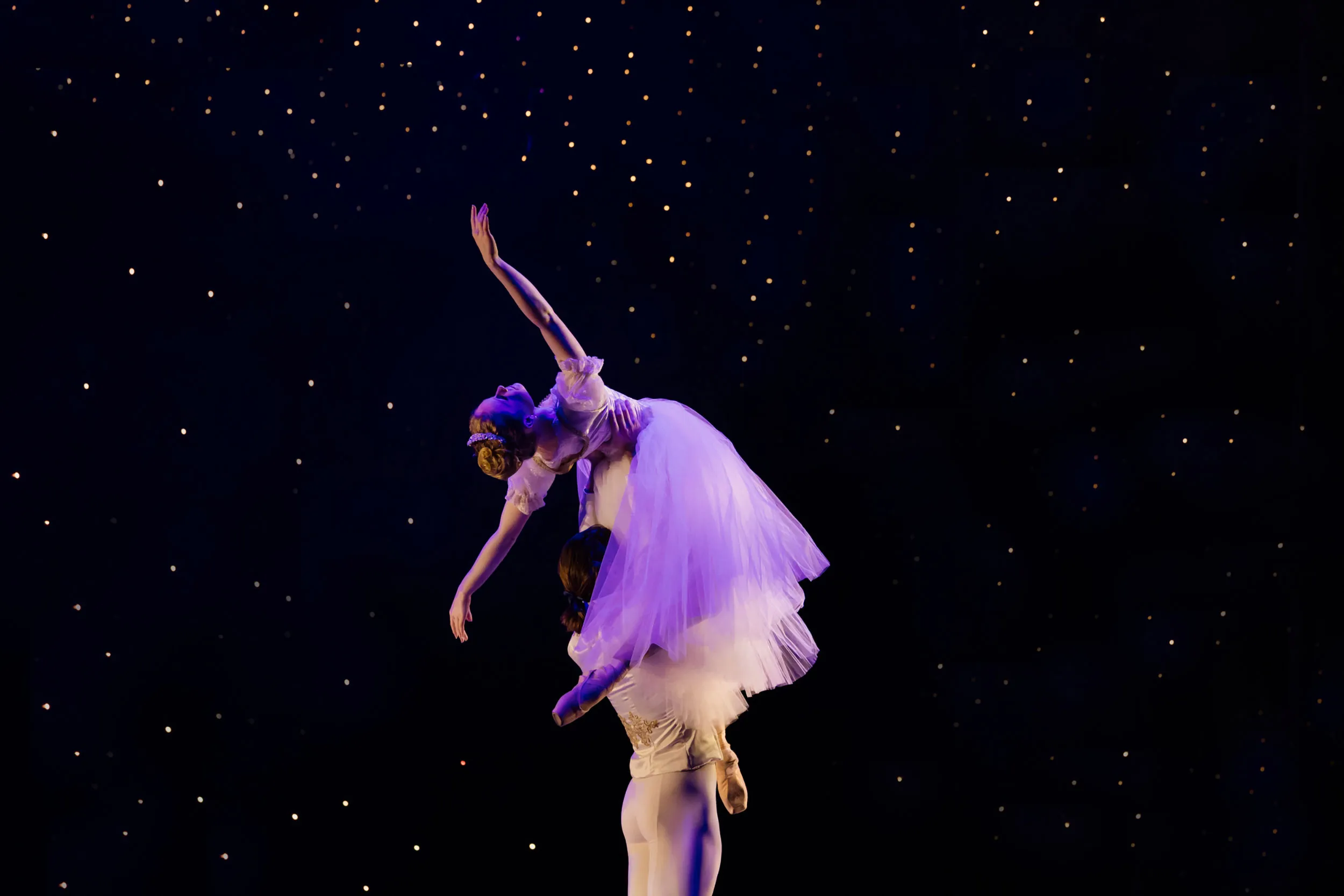 Ballet dancer lifting a female dancer in a flowing white dress against a dark starry background.