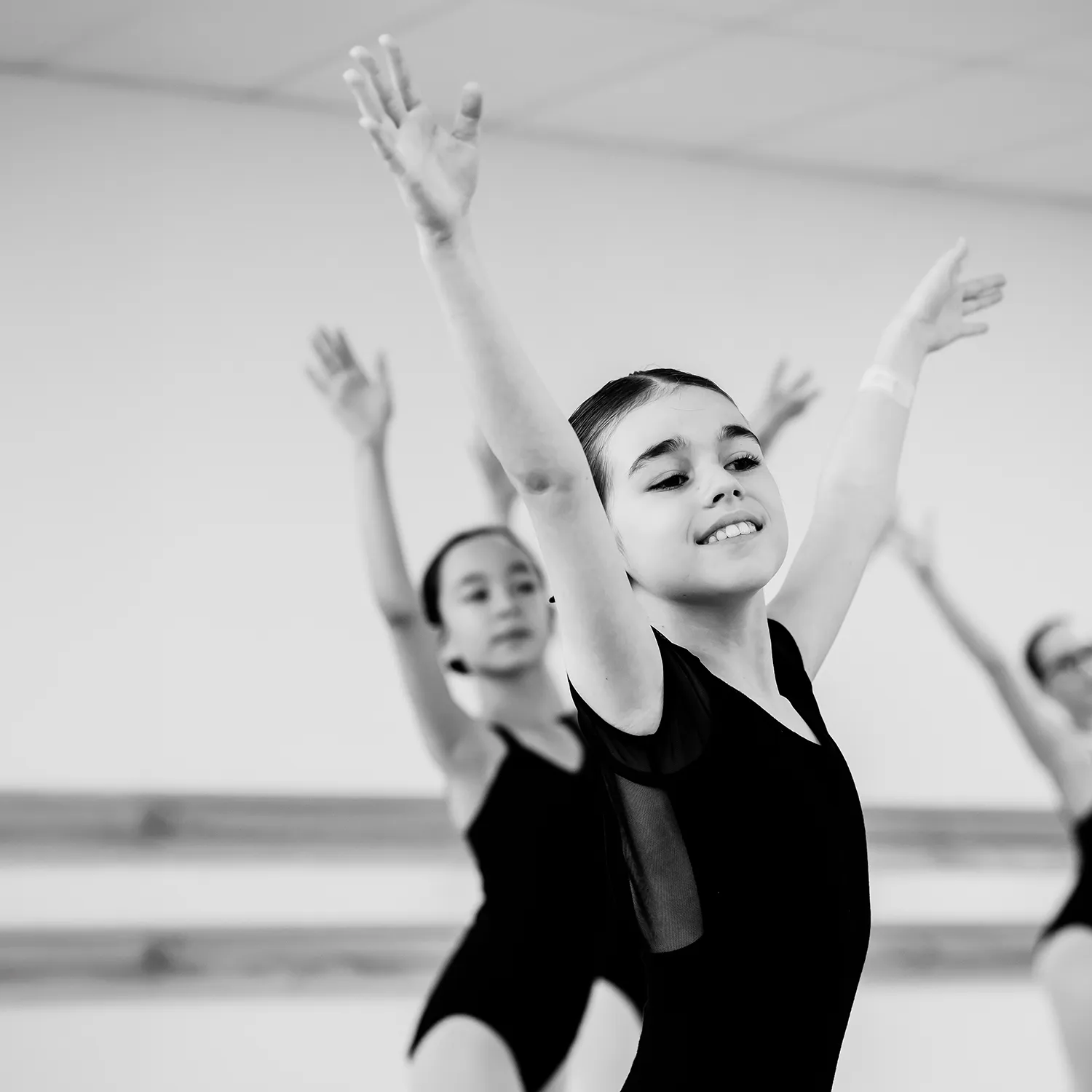 Young ballerinas in black leotards practicing with arms raised in a dance studio.