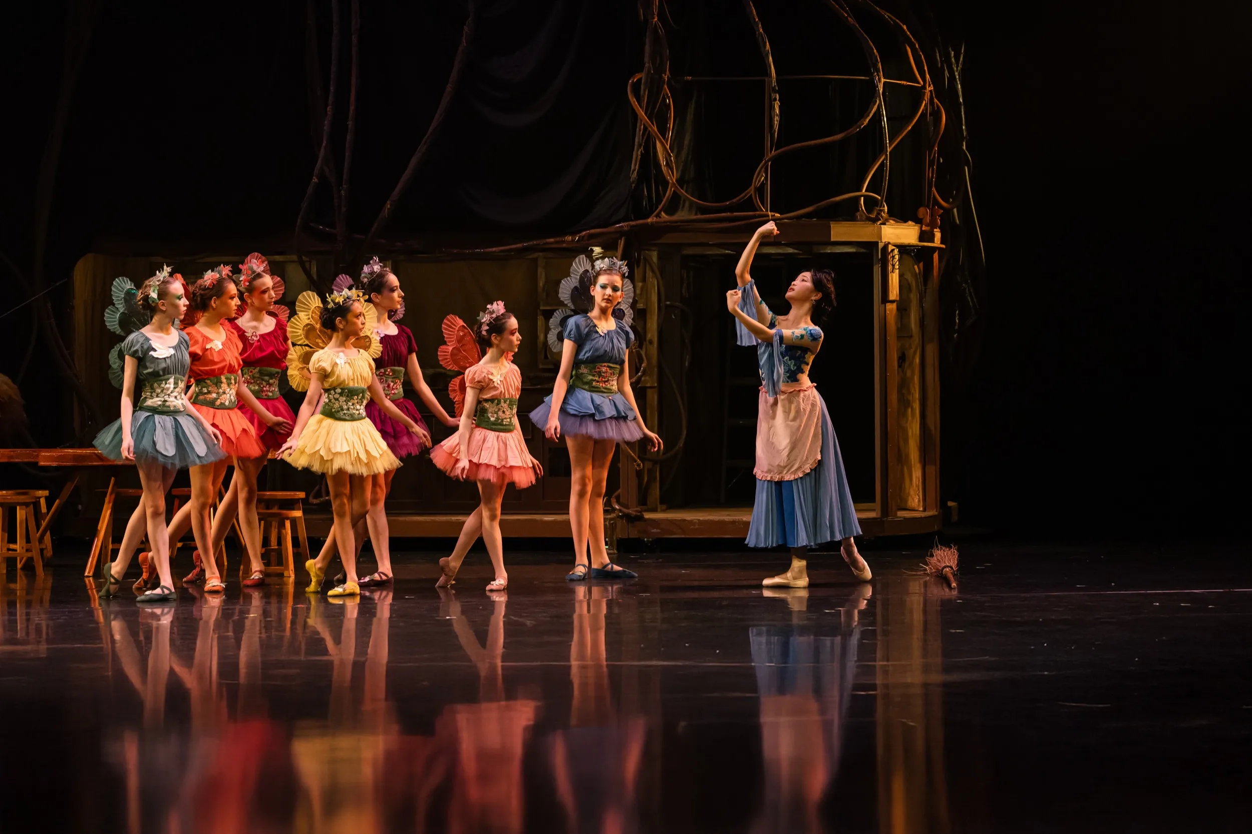 Ballet dancers in colorful tutus and flower crowns on stage with one dancer posing in a blue dress and apron near a broom.