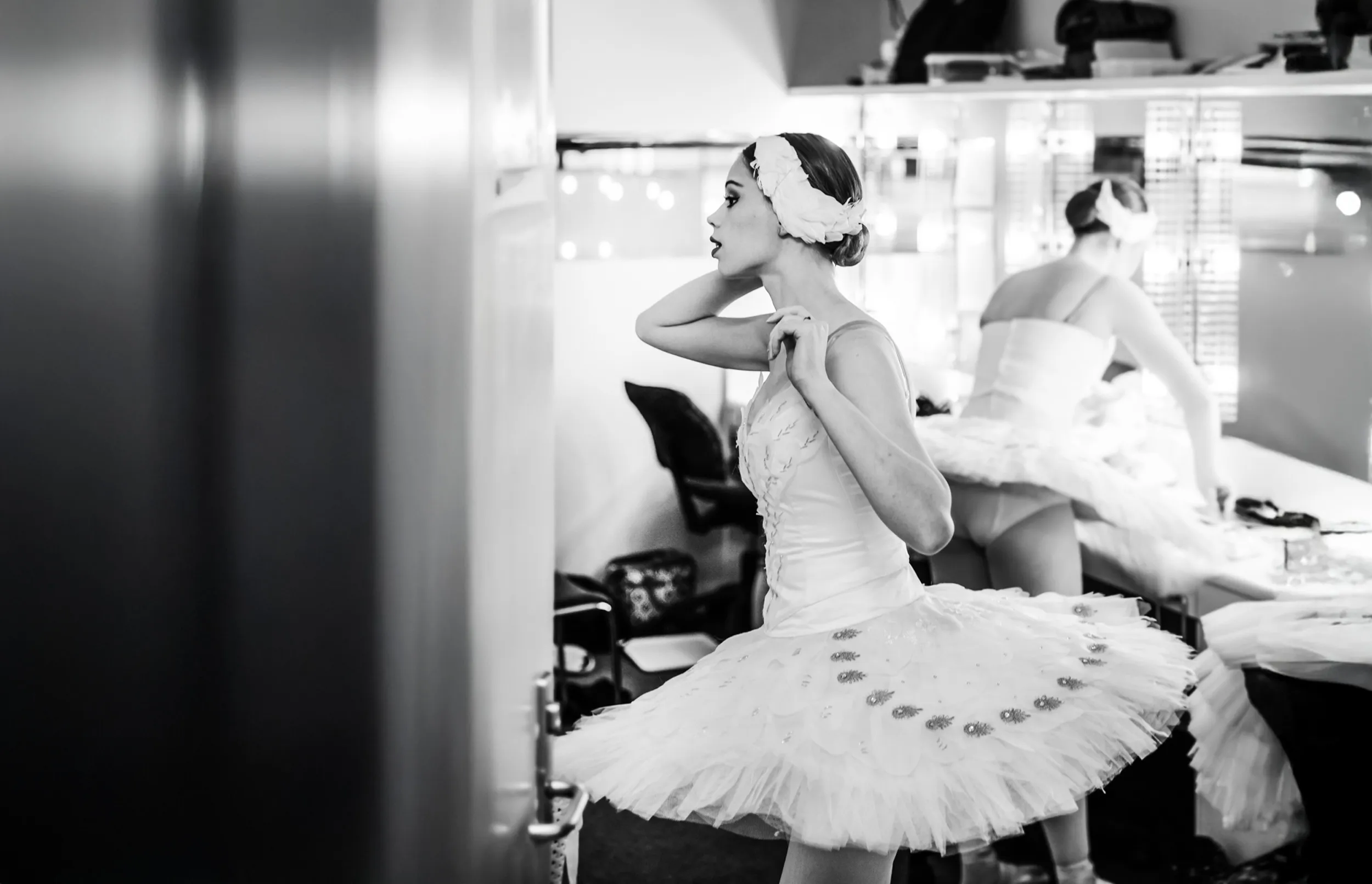 Black and white photo of a ballerina adjusting her headpiece while wearing a tutu in a dressing room with a mirror reflecting her back.