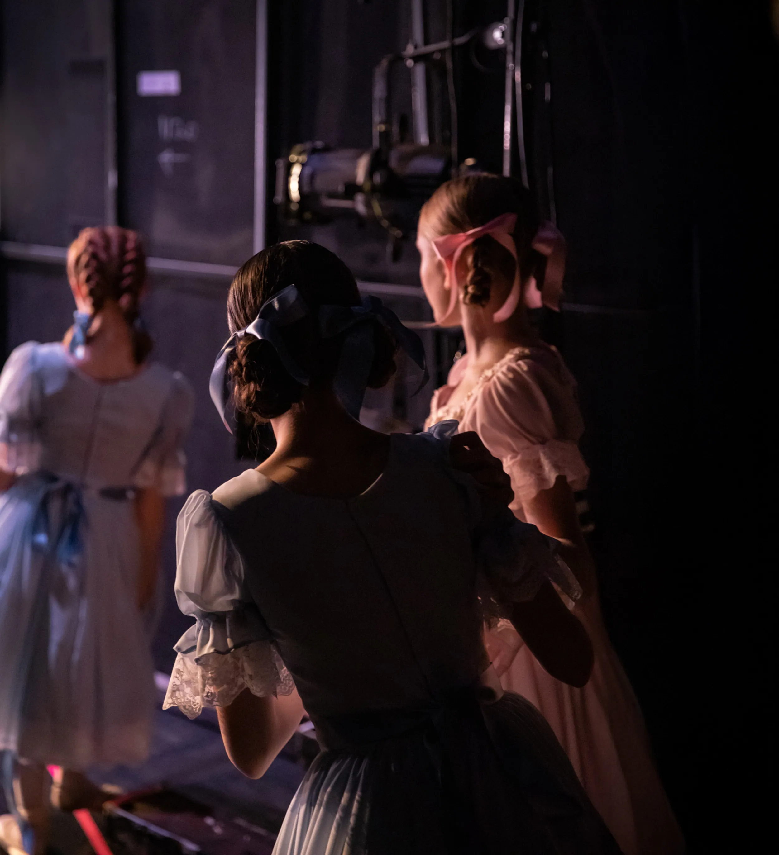 Three young girls in vintage dresses with ribboned hairstyles waiting backstage in dim lighting.