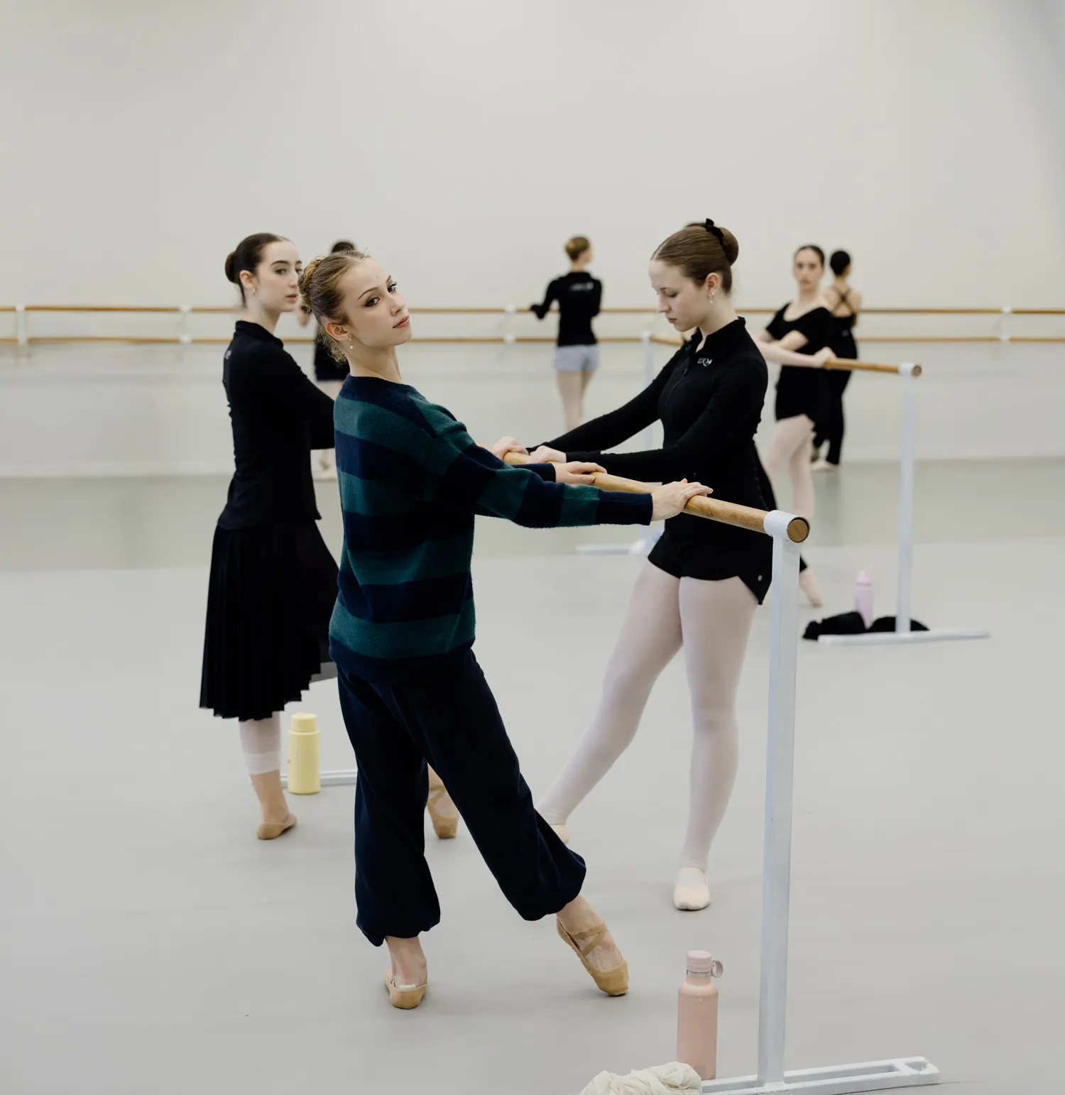 Ballet students practicing at a barre in a bright dance studio, one wearing a striped sweater and pointe shoes.