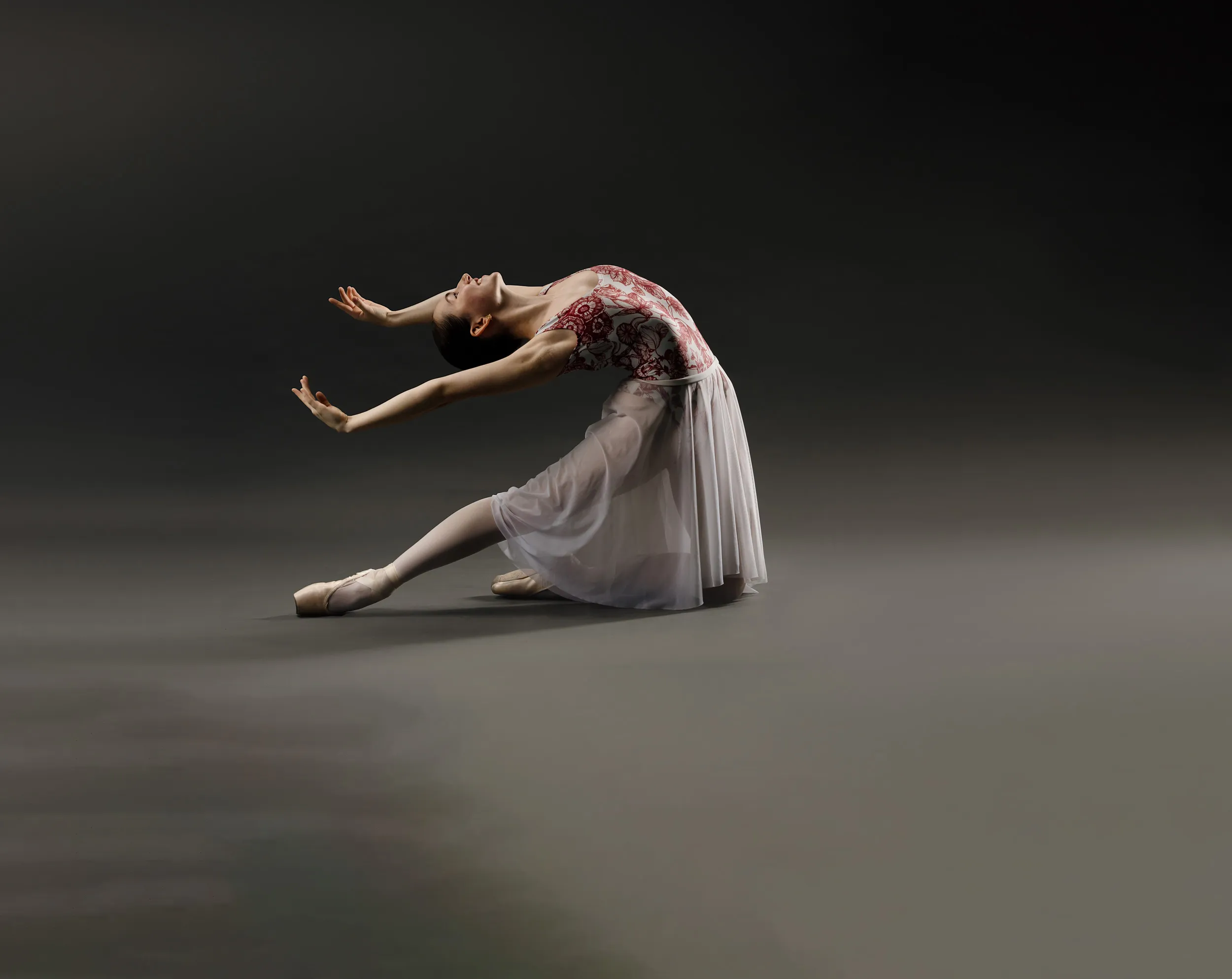 Ballet dancer in a white and red dress kneeling with back arched and arms extended gracefully against a dark background.