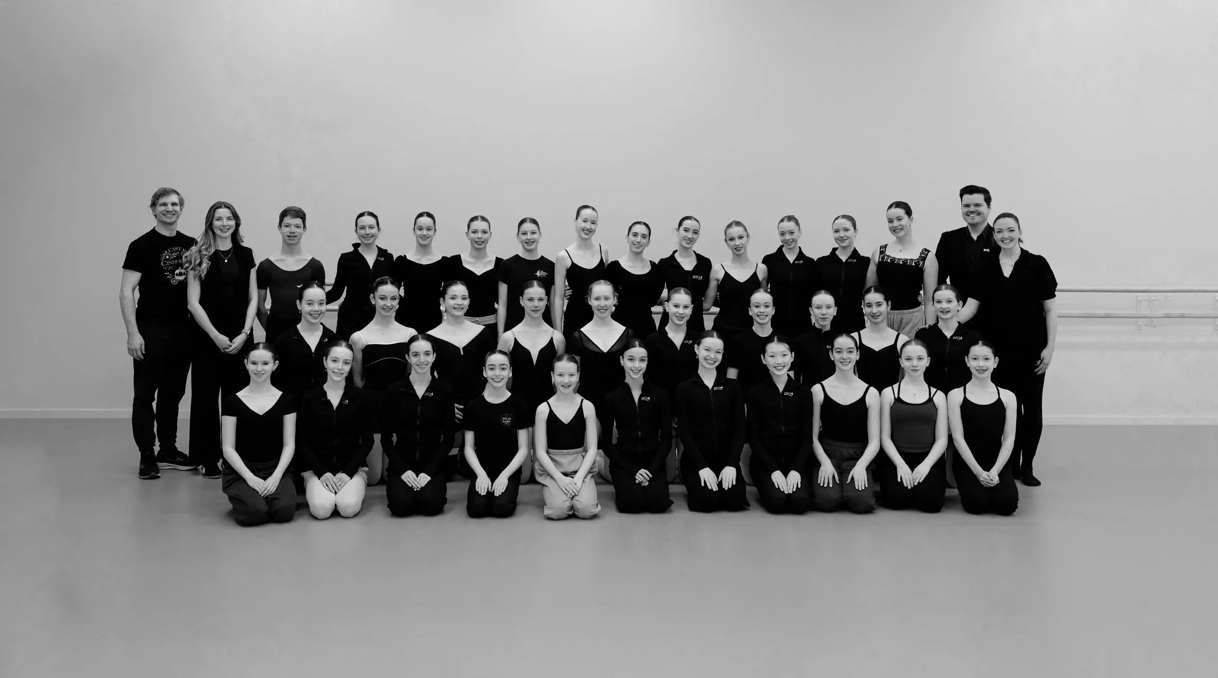 Large group of ballet dancers posing in two rows inside a dance studio with a plain wall and ballet barre.