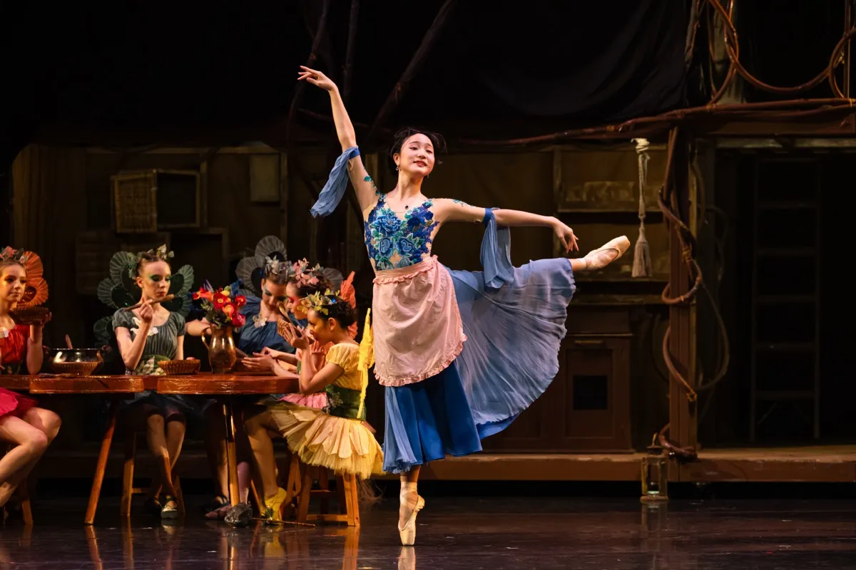 Ballet dancer in a blue floral costume and pink apron performing an arabesque on stage with seated dancers in colorful costumes behind her.