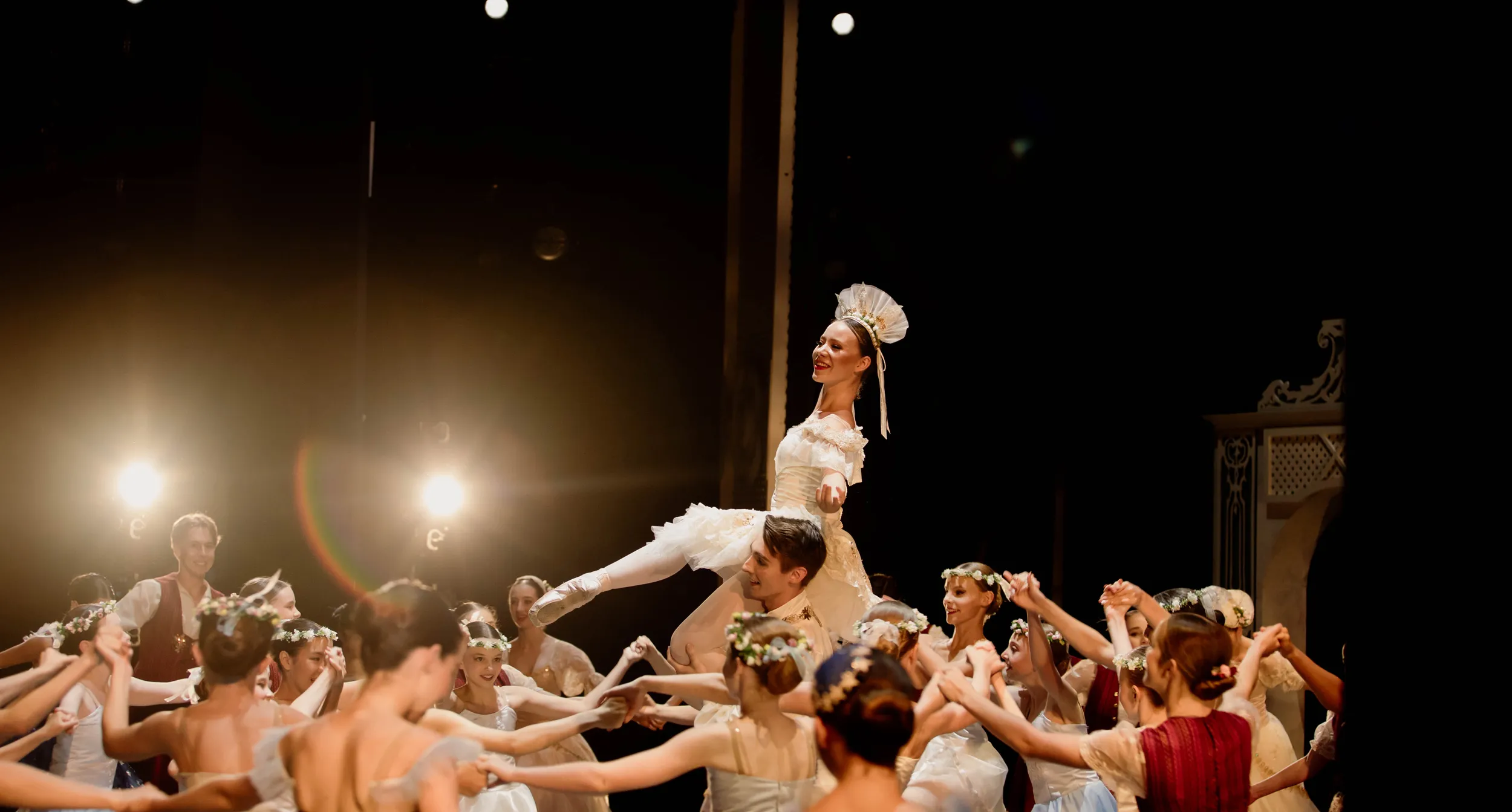 Ballet dancers in period costumes performing a group dance on stage with one dancer lifted in the center.