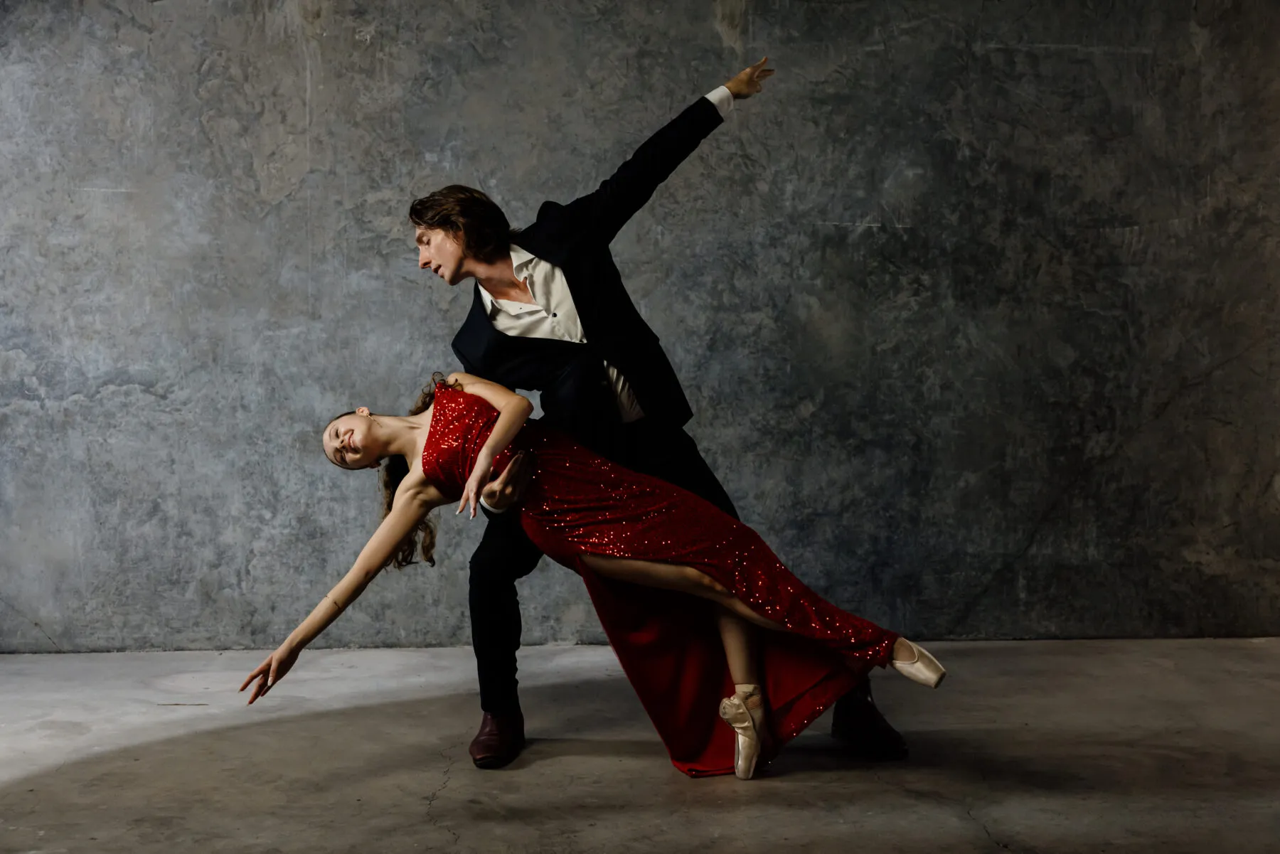 Man in black suit and woman in red sequined dress performing a dramatic dance pose against a textured gray wall.