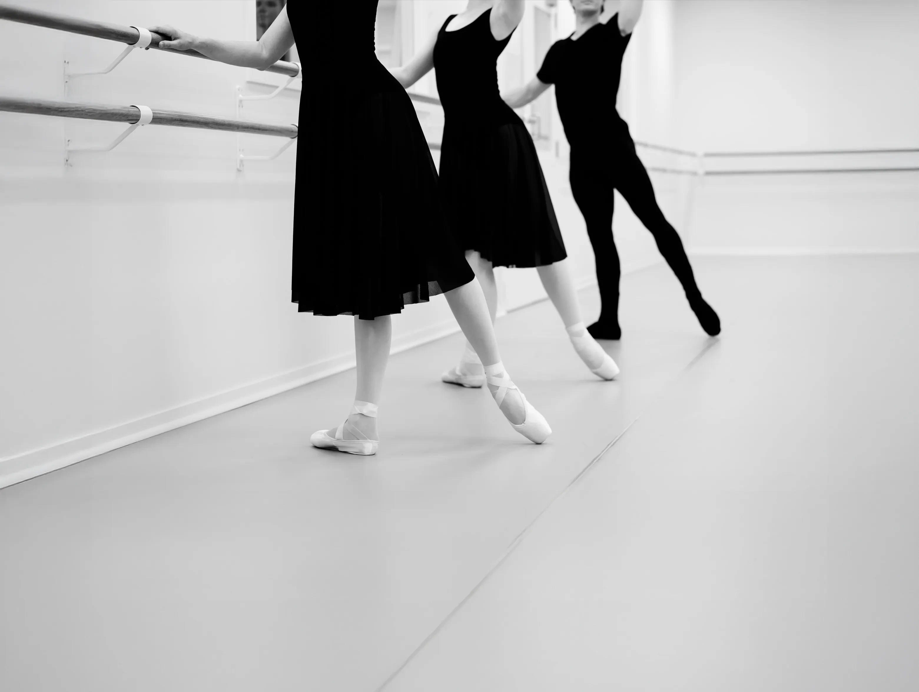 Three ballet dancers practicing at a barre in a studio, wearing black leotards and skirts, with two dancers in pointe shoes and one in ballet slippers.