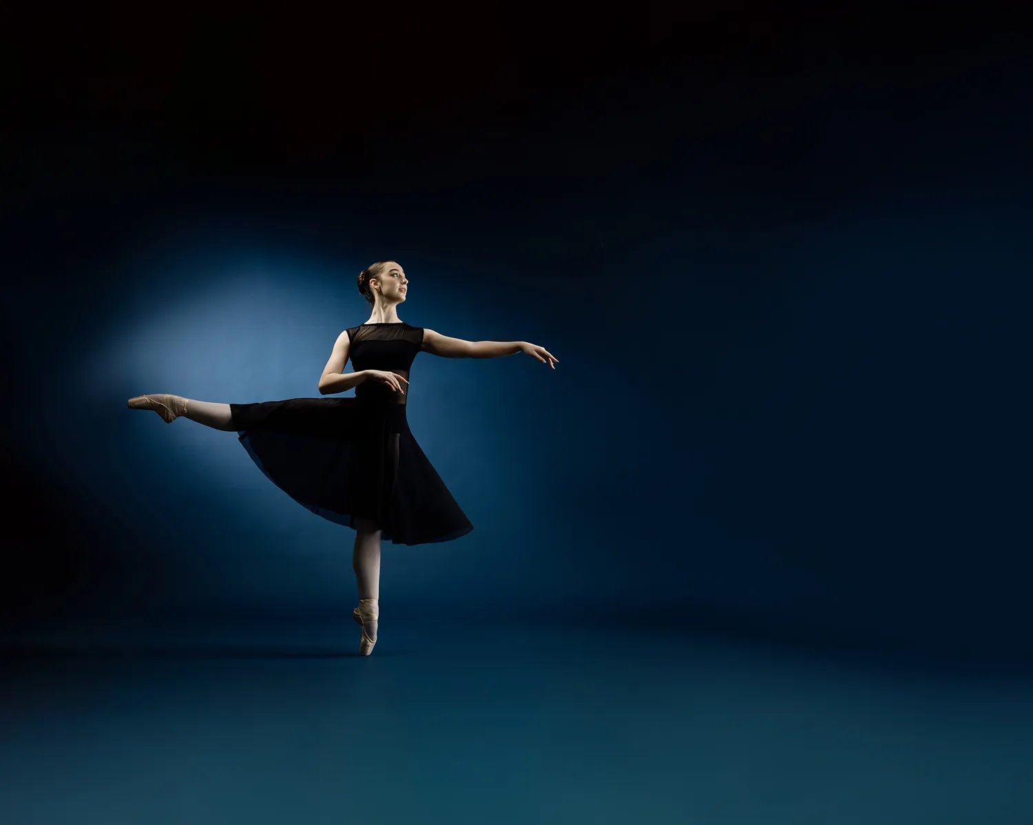 Ballet dancer in black dress performing an arabesque on pointe against a dark blue background.