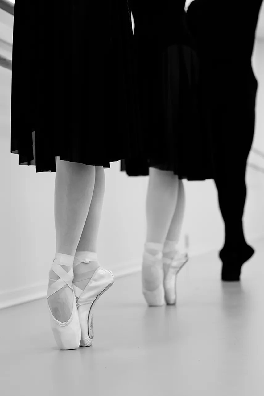 Three ballet dancers standing en pointe wearing black skirts and pointe shoes in a studio.