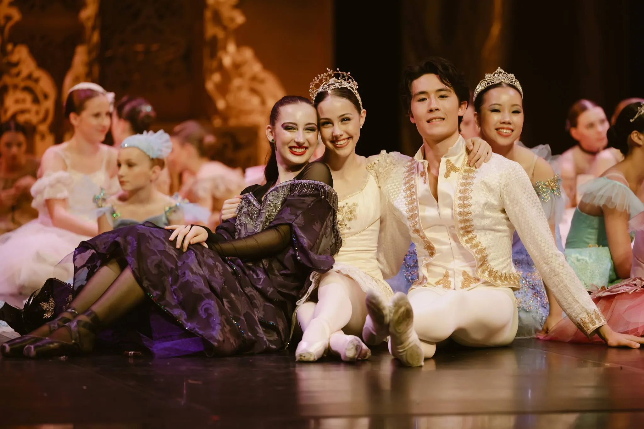 Group of ballet dancers in ornate costumes sitting on stage, smiling and posing for a photo after a performance.