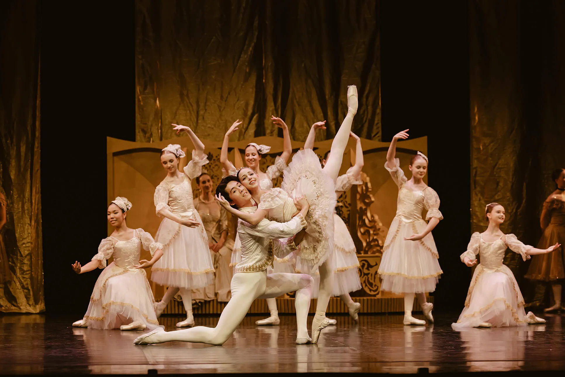 Ballet dancers in white costumes performing a classical dance with one male dancer kneeling and holding a female dancer with her leg extended high.