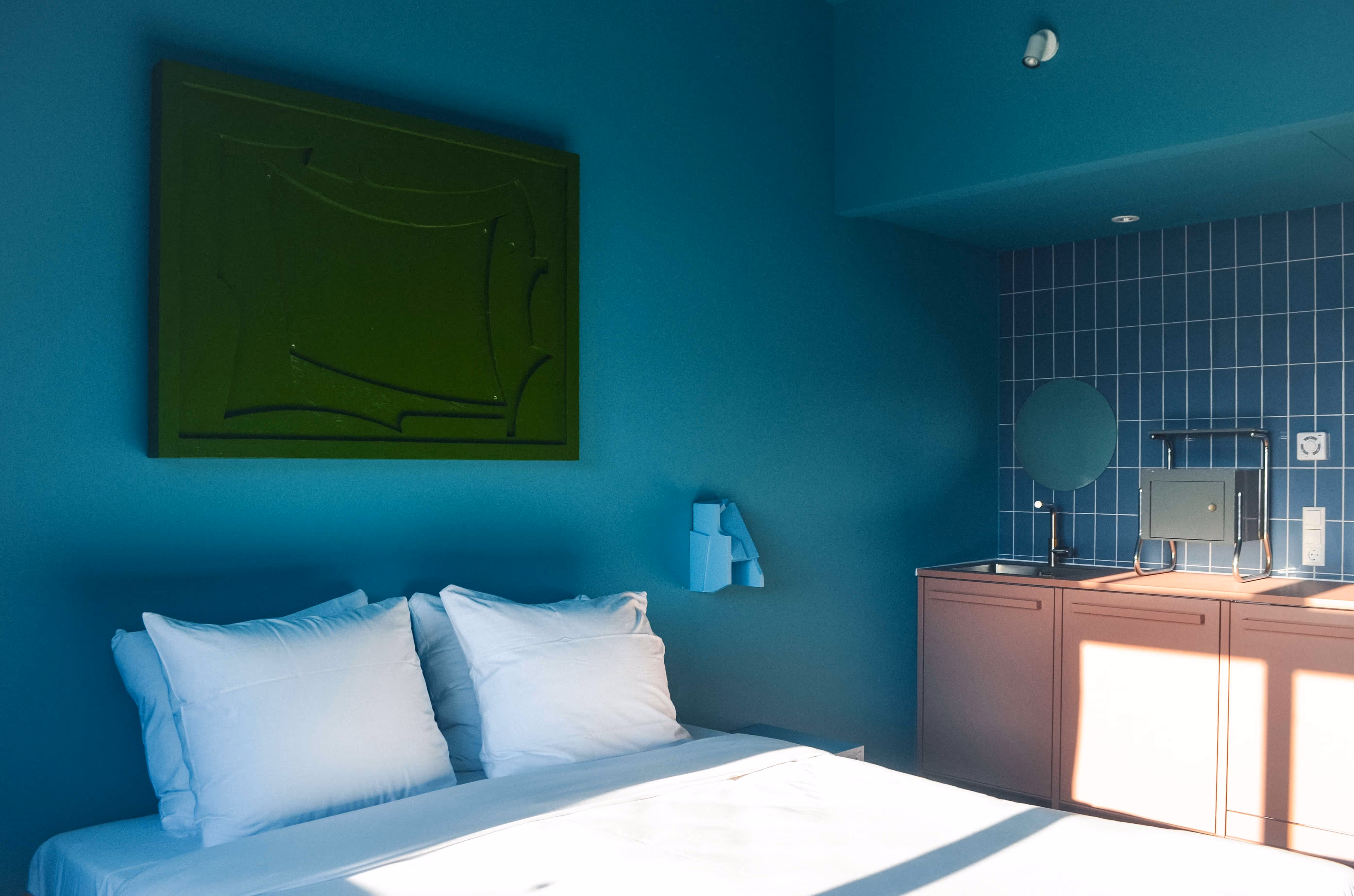 Slaapkamer met een bed met witte kussens en lichtblauwe muren, en een kleine keukenhoek met blauwe tegels en roze kasten.