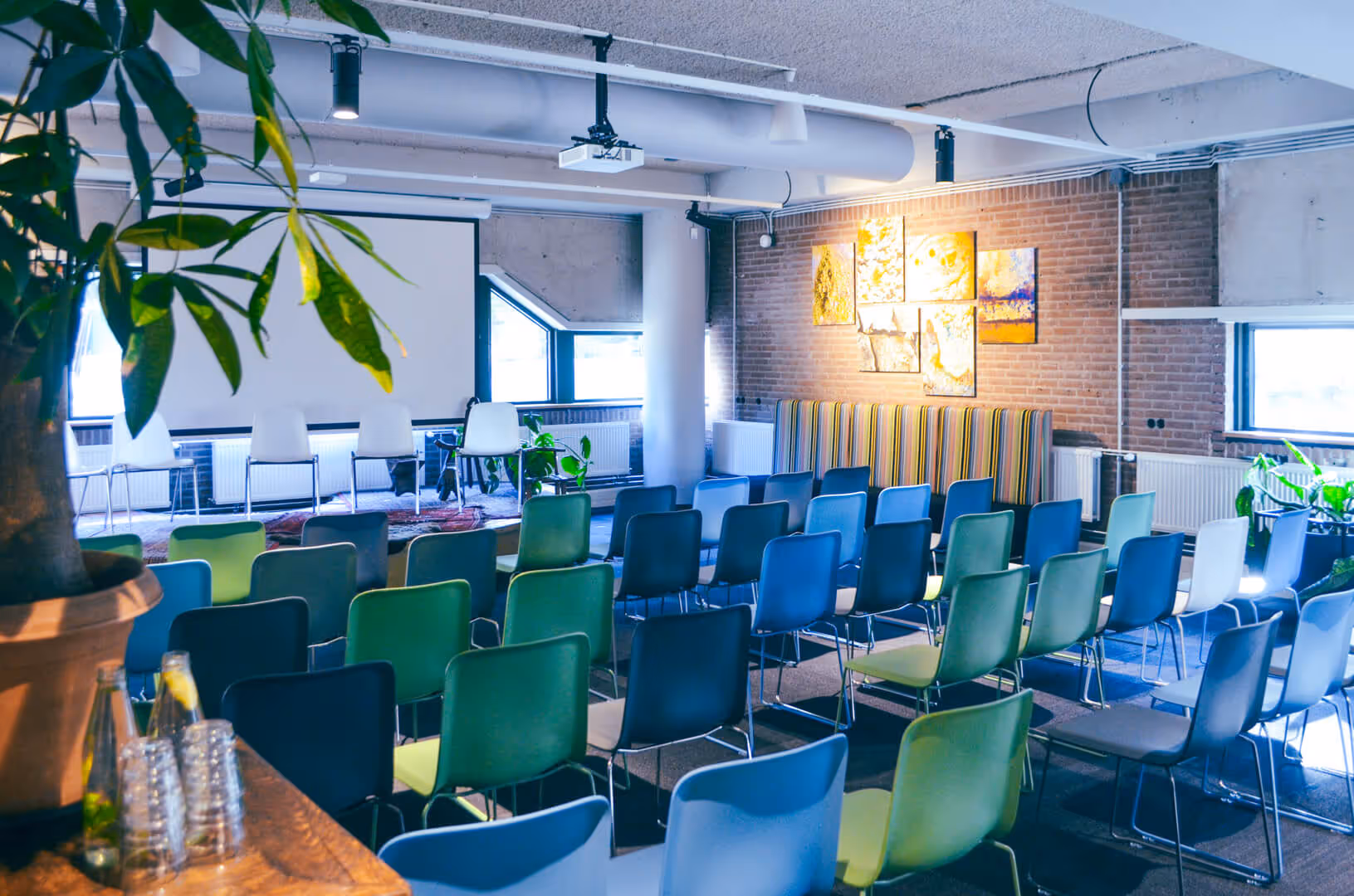 Een vergaderruimte bij De Groene Afslag met rijen gekleurde stoelen, een projector en kunst aan de muur, ingericht voor een presentatie of bijeenkomst.