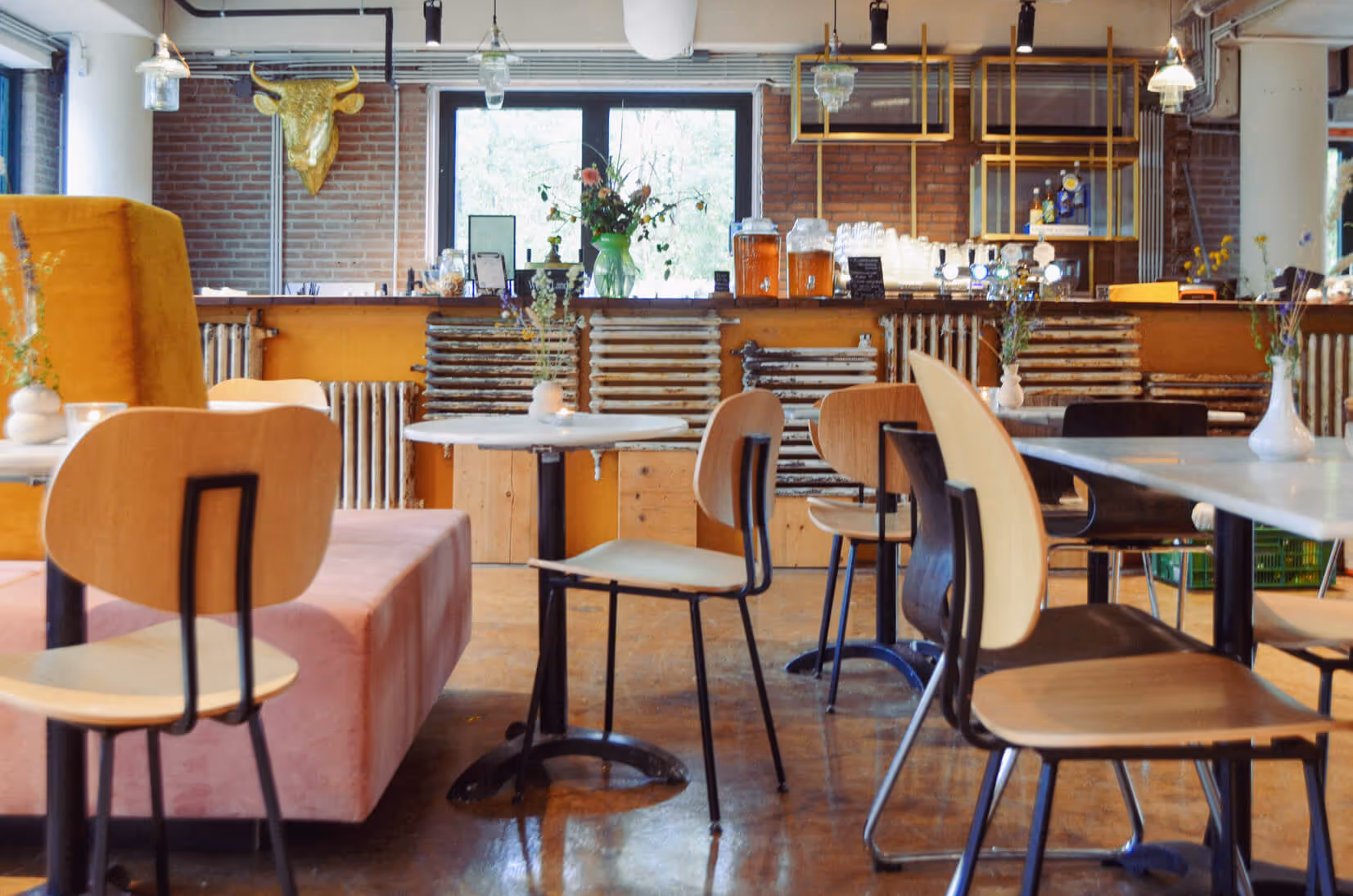 Het restaurant van De Groene Afslag met houten stoelen, roze banken en een bar van hergebruikte materialen, gevuld met bloemen en warme kleuren.